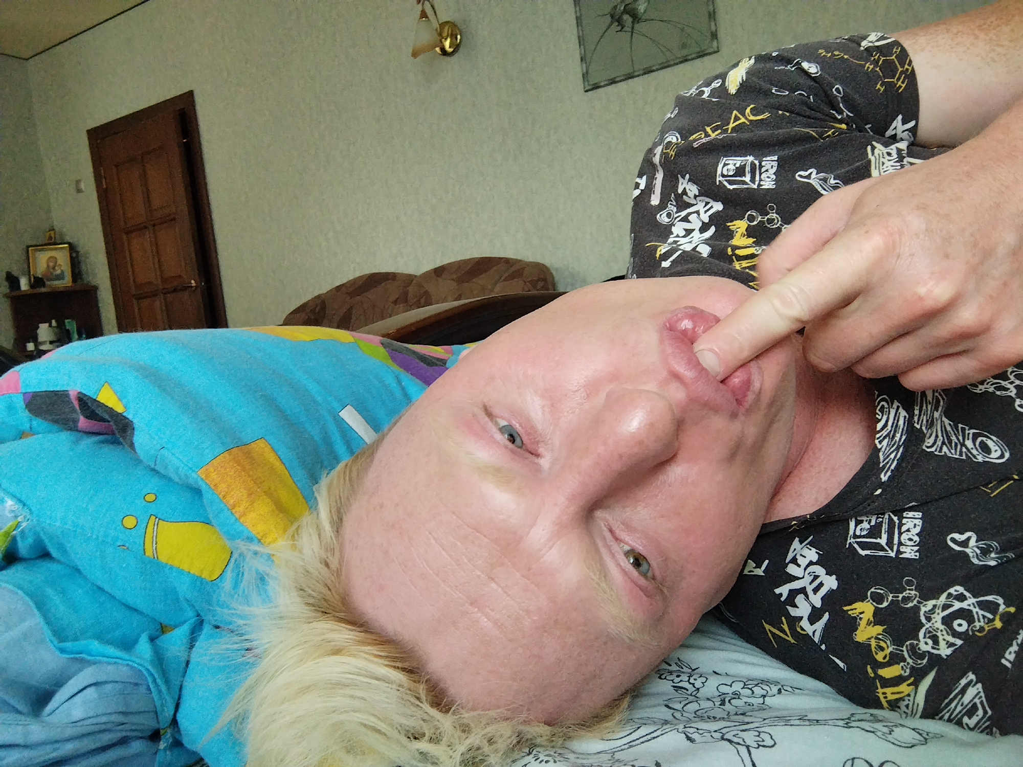 A person is lying on a bed, on their back, with their head tilted to the side. The person has blonde hair and freckled skin. They are wearing a black t-shirt with a white and yellow pattern. Their right index finger is touching their lips. The bed is covered with a blue blanket with yellow and multi-colored patterns and a white sheet with a floral pattern. In the background, there is a wooden door, a small shelf with religious icons and bottles, and a wall-mounted lamp. A brown headboard is visible behind the person. The scene appears to be indoors, in a bedroom.