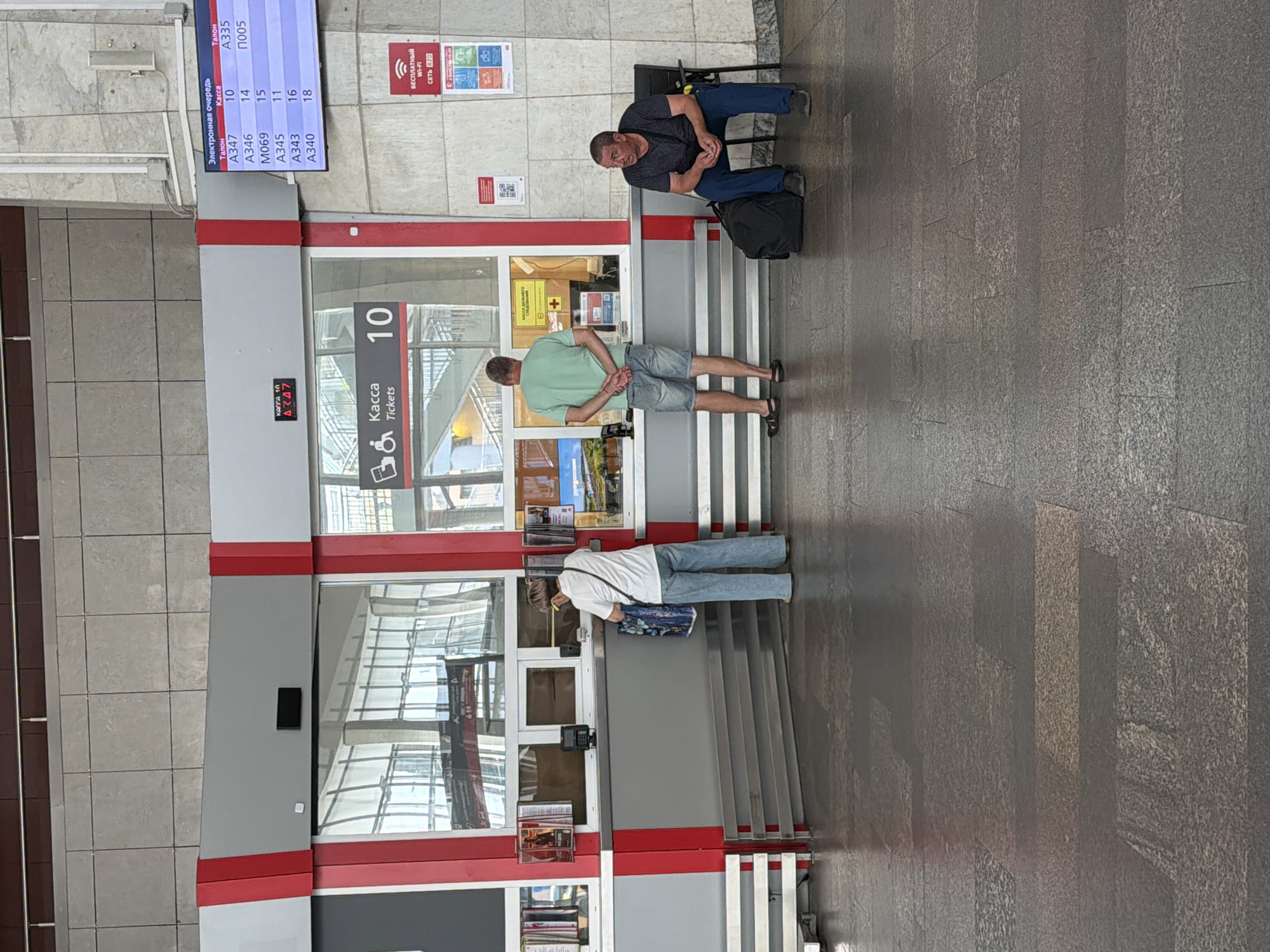 In a train station, a man in a dark t-shirt and blue pants sits on a black folding chair, looking down with his hands clasped. Beside him, a black backpack rests on the ground. In the foreground, a man in a light green t-shirt and denim shorts stands with his hands behind his back, facing away from the camera. He is looking towards a ticket counter with a digital display showing "10" and the word "Kacca" (Ticket office) in Russian. Another person, wearing a white t-shirt and blue jeans, is standing at a lower section of the ticket counter, facing away from the camera. They are holding a blue and white patterned bag.
The ticket counter is modern, with red and grey trim. Above the counter, a digital display shows a queue number "10" and other information in Russian, including "Электронная очередь" (Electronic queue) and ticket numbers like "A347" and "A335". A sign on the wall to the right, in Russian, reads "БЕСПЛАТНЫЙ WI-FI" (Free Wi-Fi). The floor is tiled with large grey and beige tiles. The overall scene suggests a busy but orderly moment at a transportation hub. The lighting indicates it's daytime, likely indoors. There are no strong emotional cues visible from the individuals.