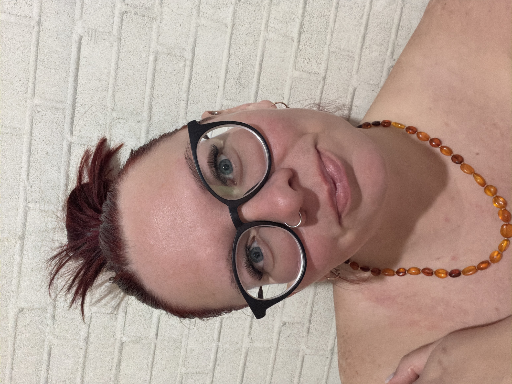 A close-up, slightly angled shot depicts a person with fair skin, auburn-red hair styled in a messy updo, and wearing black-rimmed glasses. The person has striking blue eyes accentuated by long eyelashes. A silver nose ring is visible, along with multiple earrings. They are wearing an amber-colored beaded necklace. The background is a textured, off-white surface resembling a wall. No information about Chaikovsky, Russia, is visible in the image.