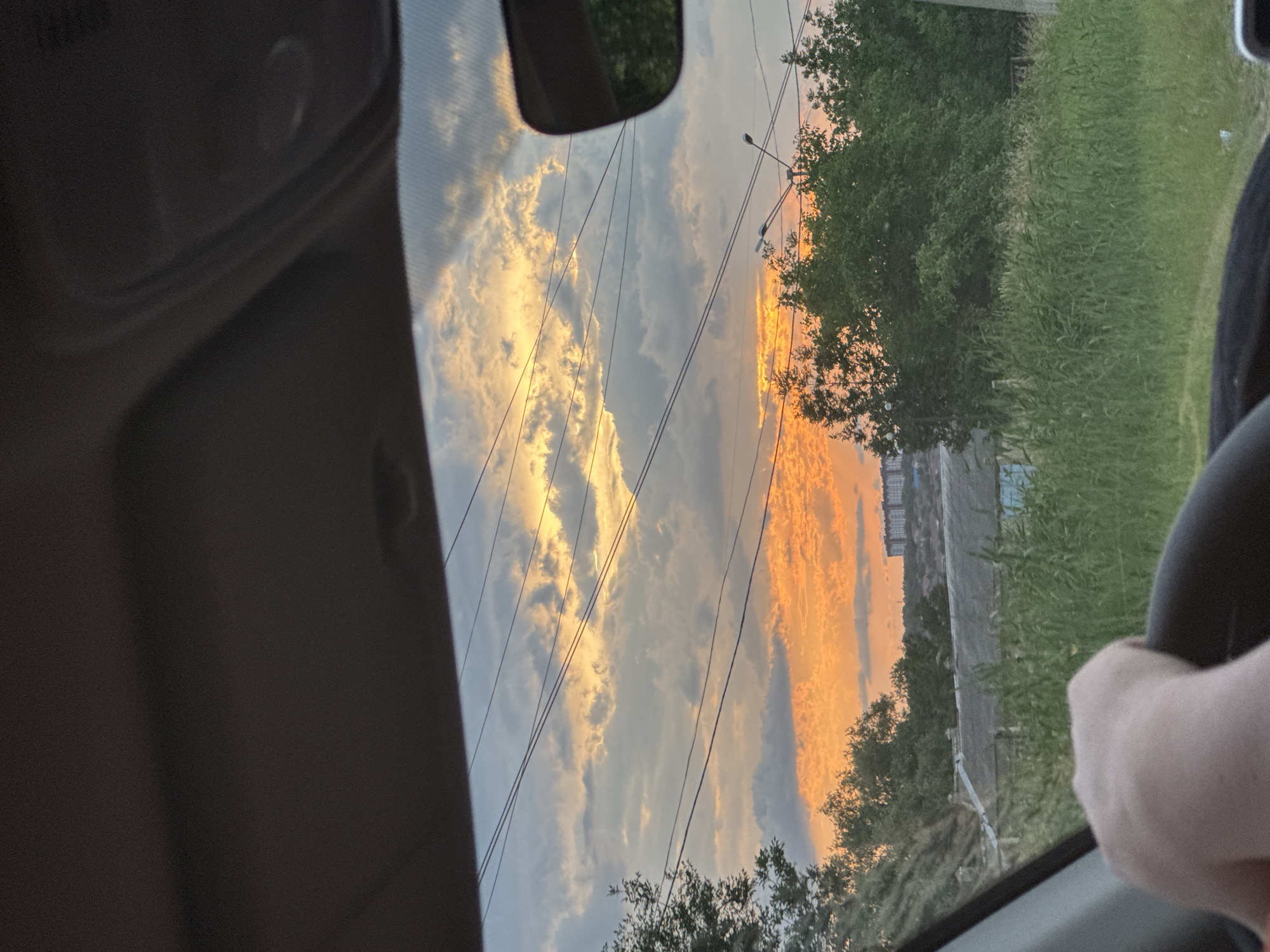 The image depicts a sunset viewed from inside a vehicle, likely a car, looking through the windshield. The sun is setting on the right side of the frame, casting a vibrant orange and yellow glow across dramatic clouds. The sky is a mix of blues, grays, and these warm sunset colors, creating a striking contrast. Several power lines crisscross the sky, adding a geometric element to the natural scene.
In the foreground, a portion of the car's interior is visible, including the dashboard on the left and a hand on the steering wheel on the right, suggesting the photo was taken while driving. Beyond the windshield, a landscape unfolds with dense green trees and tall grass. In the distance, the silhouette of buildings can be seen against the sunset, indicating an urban or semi-urban environment. The time of day is clearly evening due to the sunset. The weather appears to be partly cloudy, allowing the sun's rays to illuminate the clouds spectacularly. There is no visible text in the image. The overall mood is serene and picturesque, capturing the beauty of a sunset. The location context provided is Mikhaylovsk, Russia.