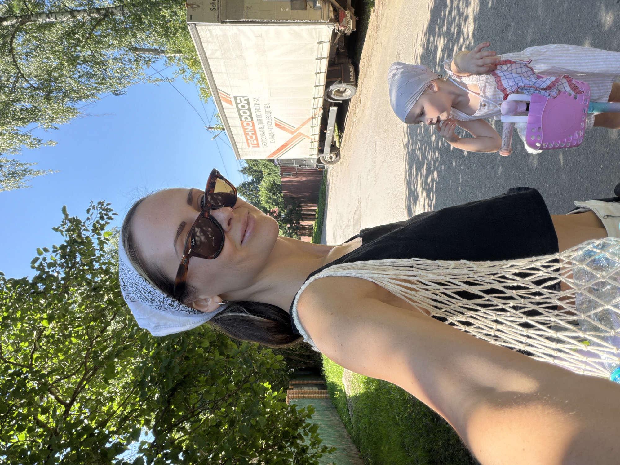 In the city of Udelnaya, Russia, on a sunny day, a woman wearing a white bandana and sunglasses takes a selfie. She is holding a white net bag containing water bottles. Next to her, a young child in a light-colored dress and a light pink bandana is sitting on a scooter with a pink basket. Behind them, a large truck with "TECHNODOOR" written on its side is parked on a paved road. Trees and a green wooden fence line the road.