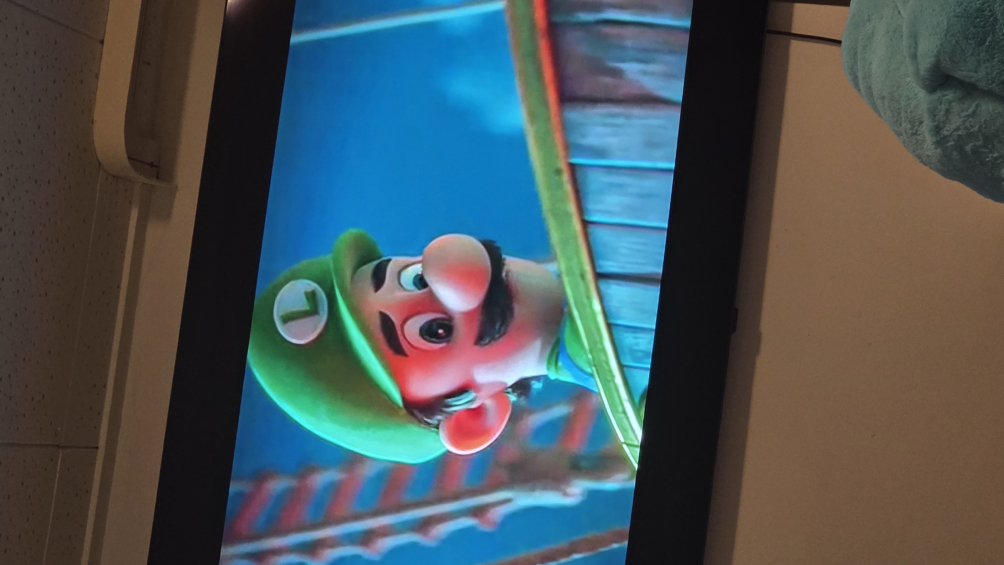 A TV screen displays Luigi from "The Super Mario Bros. Movie," positioned on an off-white wall. The screen's black frame contrasts with the colorful image of Luigi, who appears to be peering downward from a precarious position. A white pipe or fixture is visible on the left side of the screen. A turquoise object, possibly a blanket, is visible in the upper right. The location is unknown, as there is no context within the image to provide the location in the city of Zirndorf, Germany.