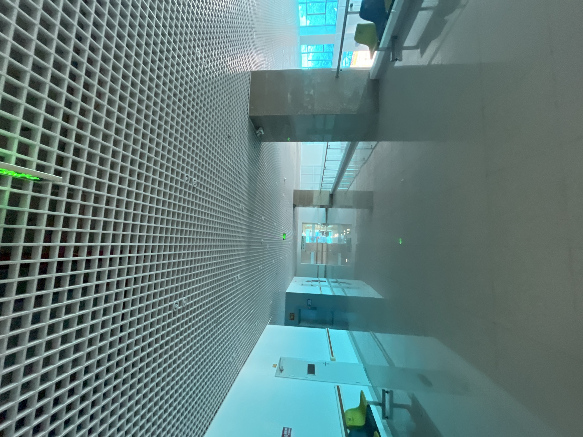 The image shows a corridor or hallway in what appears to be an airport terminal or a similar transportation hub. The grid-like structure on the left suggests a wall or ceiling design feature. The right side reveals a smooth, reflective surface, likely a polished floor. The perspective is from a high angle, looking down the length of the corridor. There are glimpses of seating areas with blue and green chairs and other infrastructure elements within the building. The location is in Nha Trang Ward, Vietnam.