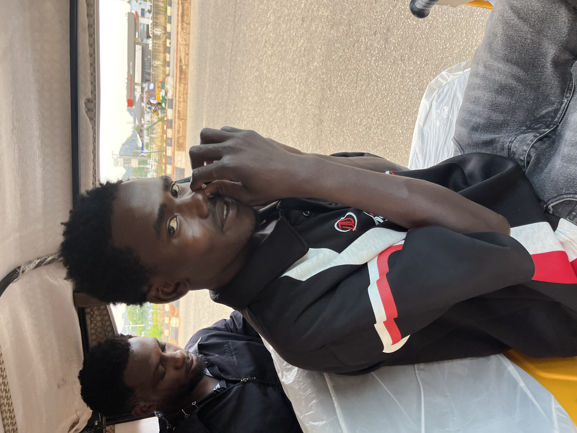 Two young Black men are seated inside a tuk-tuk on a paved road in Bukuru, Nigeria. The man in the foreground, facing the camera, is wearing a black polo shirt with red and white accents on the sleeves and collar. He has his hands raised to his face, touching his nose. The man behind him is wearing a black zipped jacket. The interior of the tuk-tuk is visible, with white plastic sheeting and a patterned fabric lining the roof. In the background, a gas station with red and white signage is visible across the road, along with other vehicles, including a motorcycle with two people. The sky is overcast.