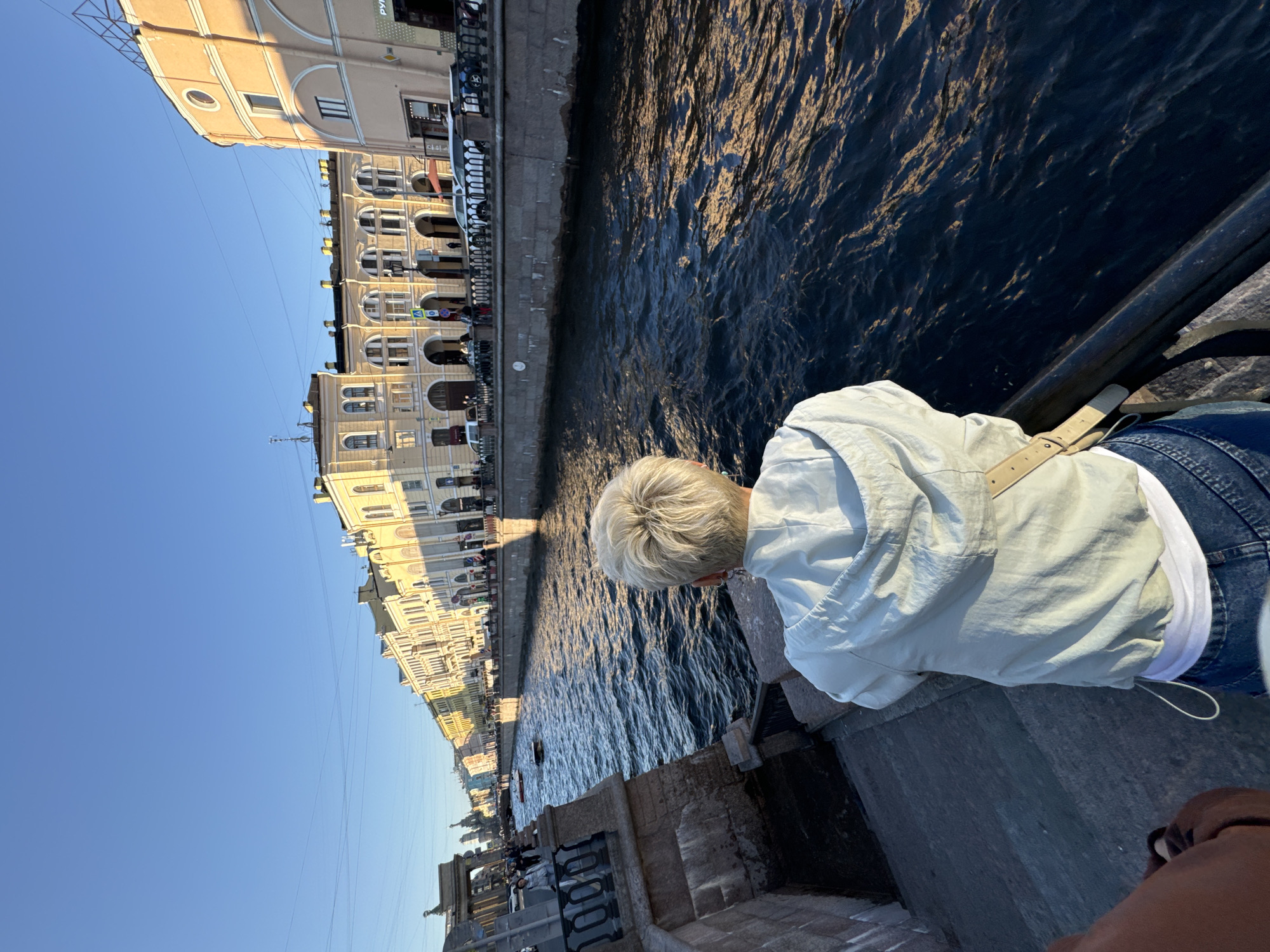In Saint Petersburg, Russia, a person with short, blonde hair is seen from behind, facing a canal. The person is wearing a light-colored hooded jacket, a white undershirt, and blue jeans. They are standing on a stone embankment, possibly a bridge or a walkway, with a dark railing in the foreground. The canal water is dark blue and reflects the warm golden light of the setting sun, creating ripples and highlights on its surface. Across the canal, a row of historic buildings with yellow facades and arched windows lines the embankment. These buildings exhibit architectural features common in Saint Petersburg, such as classical detailing and mansard roofs. The sky is clear and blue, indicating a sunny day. The overall scene captures a serene moment along a scenic canal in the city.