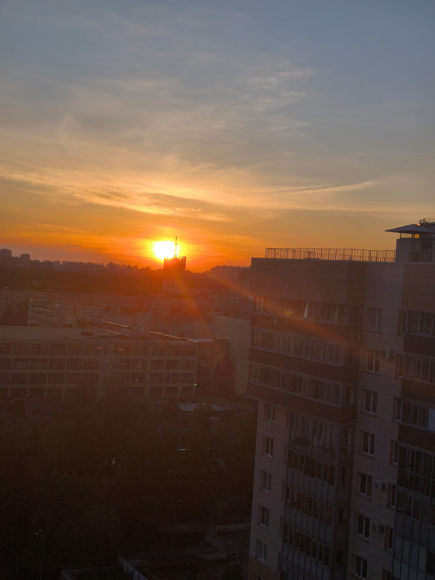 The image shows a cityscape at sunset. The sun is setting behind a building with a tall spire, casting a bright orange glow across the sky and illuminating the surrounding buildings. The sky transitions from orange near the horizon to a lighter blue at the top. In the foreground, there is a multi-story building with multiple windows. Below the buildings is a dark area with what appears to be trees and some structures. The location is unknown, but it is a city with a mix of residential and potentially commercial buildings.