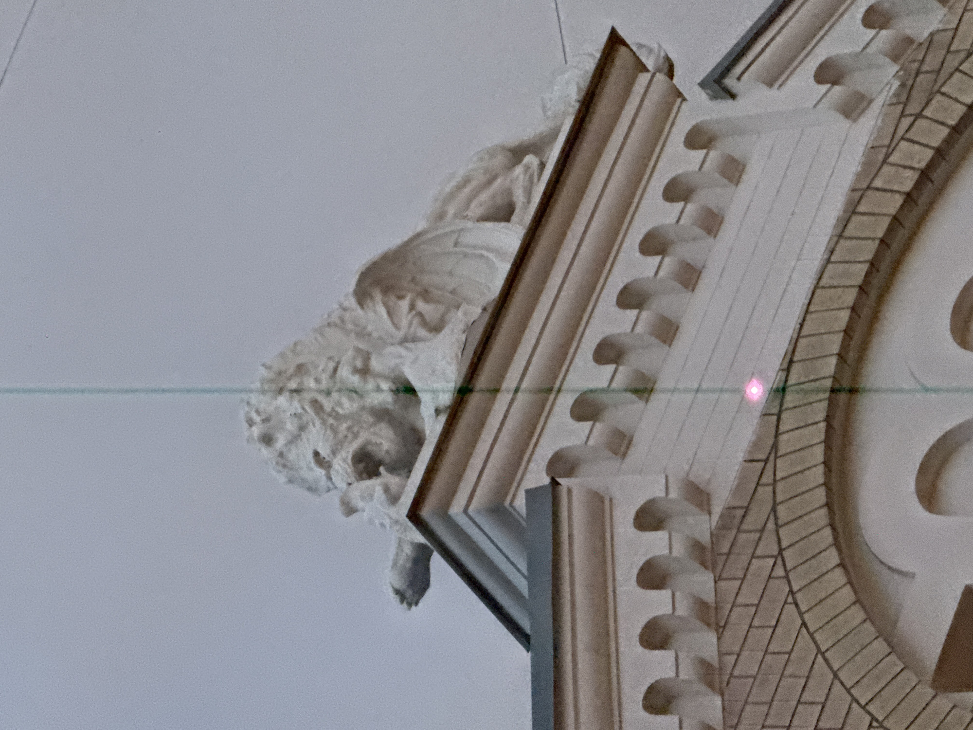 The image shows a close-up, upward view of architectural details from what appears to be a building's interior in Moscow, Russia. A carved, off-white angel figure is positioned to the left, partially obscured by ornate moldings. The moldings are predominantly white with hints of beige and are adorned with patterns and a brick-like texture on the curved section. A thin green line with a small pink dot cuts across the middle of the frame horizontally. The background is a solid light gray, suggesting the ceiling. The overall setting conveys a sense of classical architecture.