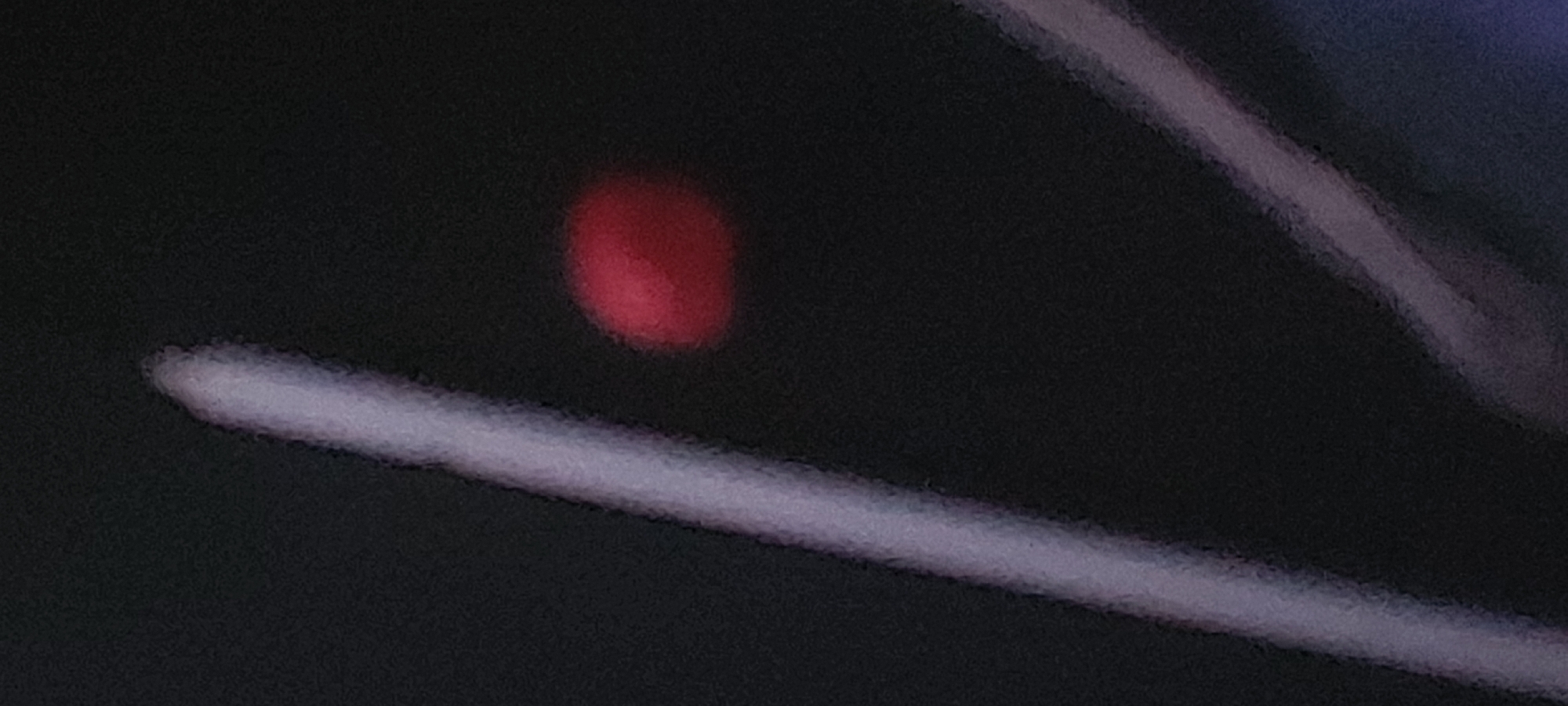 The image features a close-up shot of a dark surface, possibly part of a dashboard or electronic device, with a blurred red circular light indicator. A light-colored, elongated object resembling a wire or thin tube is positioned diagonally across the lower portion of the frame. The focus is soft, creating a sense of obscurity, and the ambient light is low, contributing to the overall dark and indistinct quality of the image. There is no identifiable information that can be associated with Tashkent, Uzbekistan.