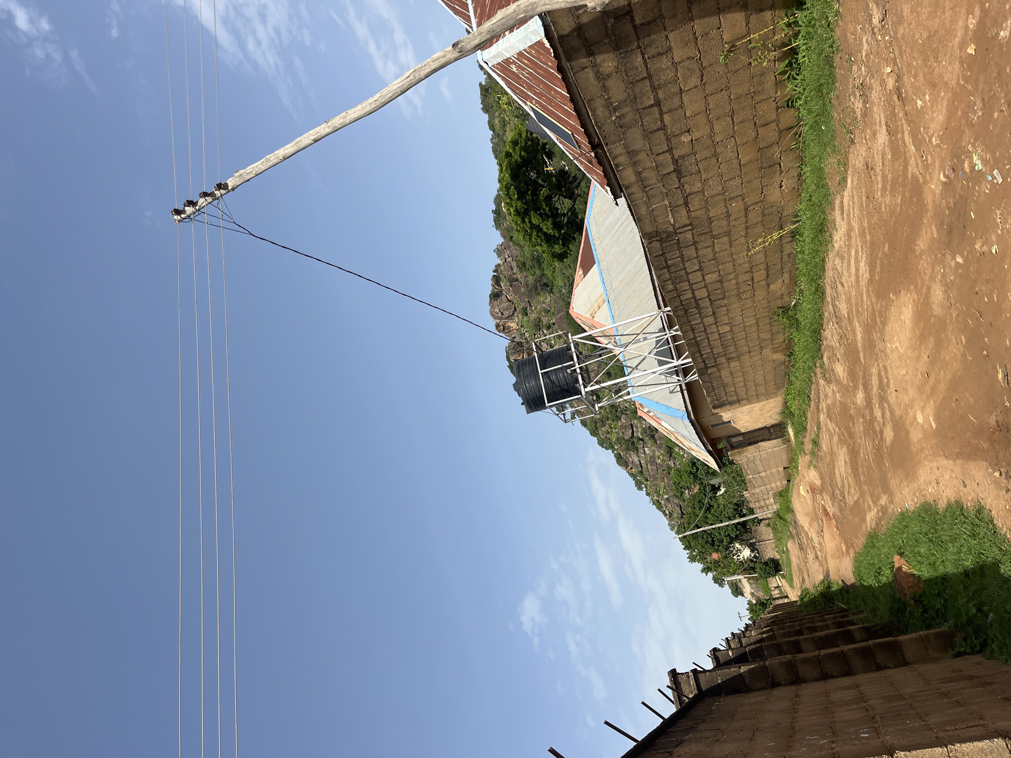 The image shows a dirt road in Bukuru, Nigeria, flanked by a high brick wall on the left and a lower brick wall on the right. Several buildings with corrugated metal roofs are visible beyond the lower wall, and a water tank on a metal stand is perched on top of one of them. A wooden utility pole with power lines extends diagonally across the frame from the lower right to the upper left. In the background, a rocky, tree-covered hillside rises under a clear blue sky with a few scattered clouds. The scene appears to be a residential or semi-developed area.