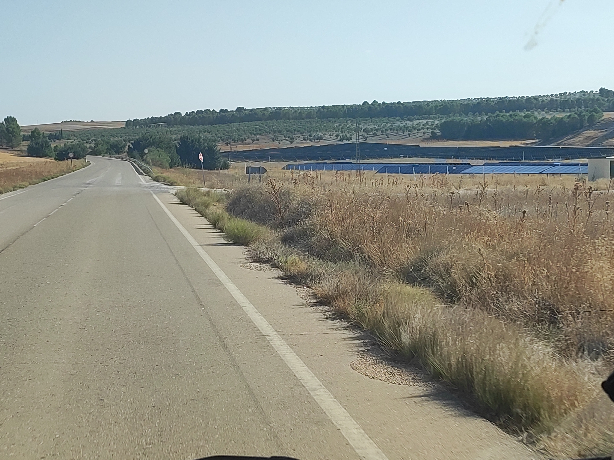The image captures a road leading through a rural landscape in the vicinity of Huelves, Spain. The road, paved with asphalt and marked with a white line, curves gently to the left. On the right side of the road, dry, tall grasses and sparse vegetation line the shoulder, transitioning into a field containing a large solar farm. Rows of blue solar panels are clearly visible within the solar farm, stretching across the terrain. In the distance, a dense line of trees, possibly an olive grove, runs along a slightly elevated horizon. The sky is clear and bright, indicating a sunny day. There are no people or vehicles immediately visible in the frame. The overall scene depicts a semi-arid agricultural and energy production area.