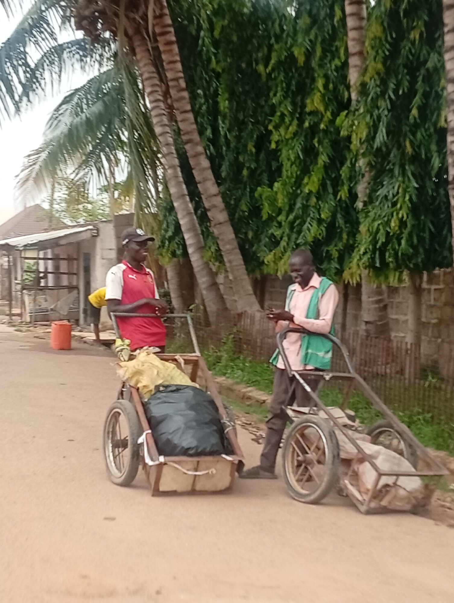 The image shows two men on a street in what appears to be Sabon Tasha, Nigeria. One man is wearing a red shirt, black cap, and is standing behind a homemade cart containing a black garbage bag and other materials. The second man is wearing a light pink shirt and a green vest. He is holding a mobile phone and standing behind a similar cart loaded with what appears to be waste. Both men are facing each other and smiling. In the background, there are palm trees, tall bushes, and a building with a corrugated metal roof. An orange trash bin is visible in the background. The street is unpaved and made of dirt.