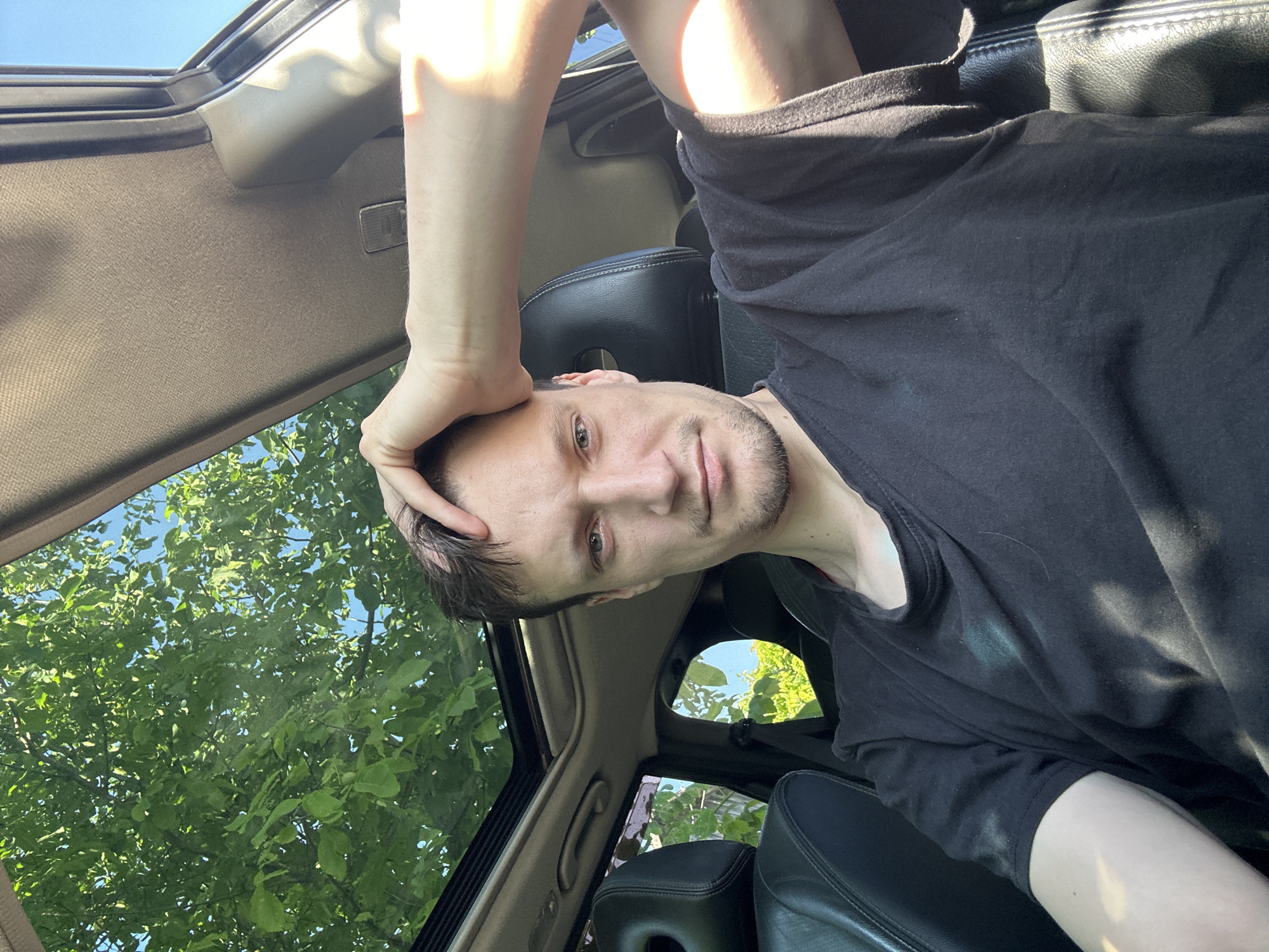 In Shatava, Ukraine, a young man with light brown hair and a short beard sits in the driver's seat of a car, with his right hand resting on his head. He is wearing a black t-shirt. The car's sunroof is open, revealing green foliage and a clear blue sky above. Sunlight streams into the car, casting shadows on the interior and the man's clothing. The car's black leather seats are visible in the foreground.