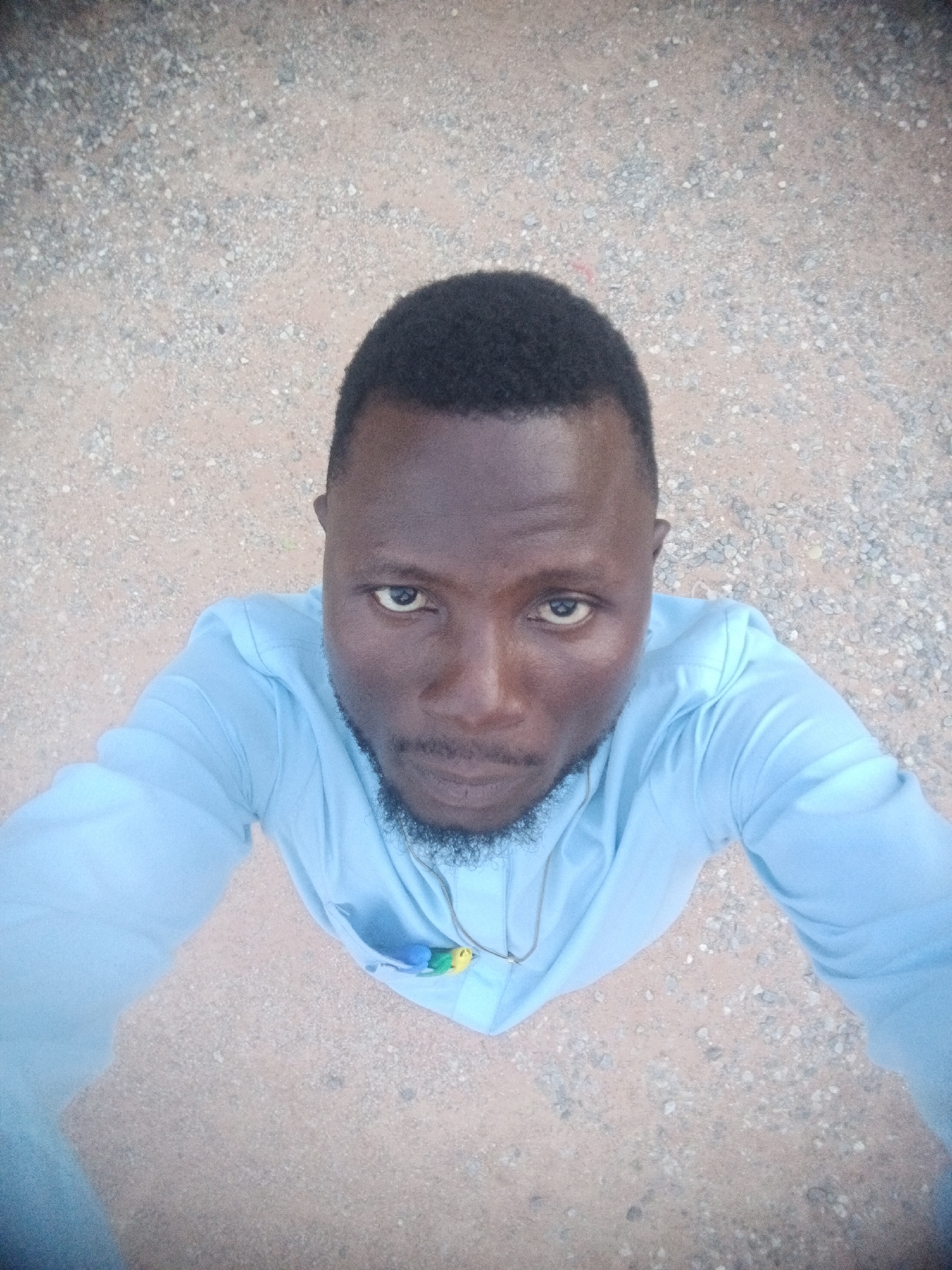 The media file is a selfie taken from a high angle, looking down at a man. The man is wearing a light blue collared shirt and has short black hair and a beard. He is looking directly at the camera with a neutral expression. A necklace with a small, colorful pendant is visible around his neck. The background is an outdoor setting with a gravelly, sandy ground. The lighting suggests it is daytime. Based on the provided location context, the scene is likely in Jalingo, Nigeria.