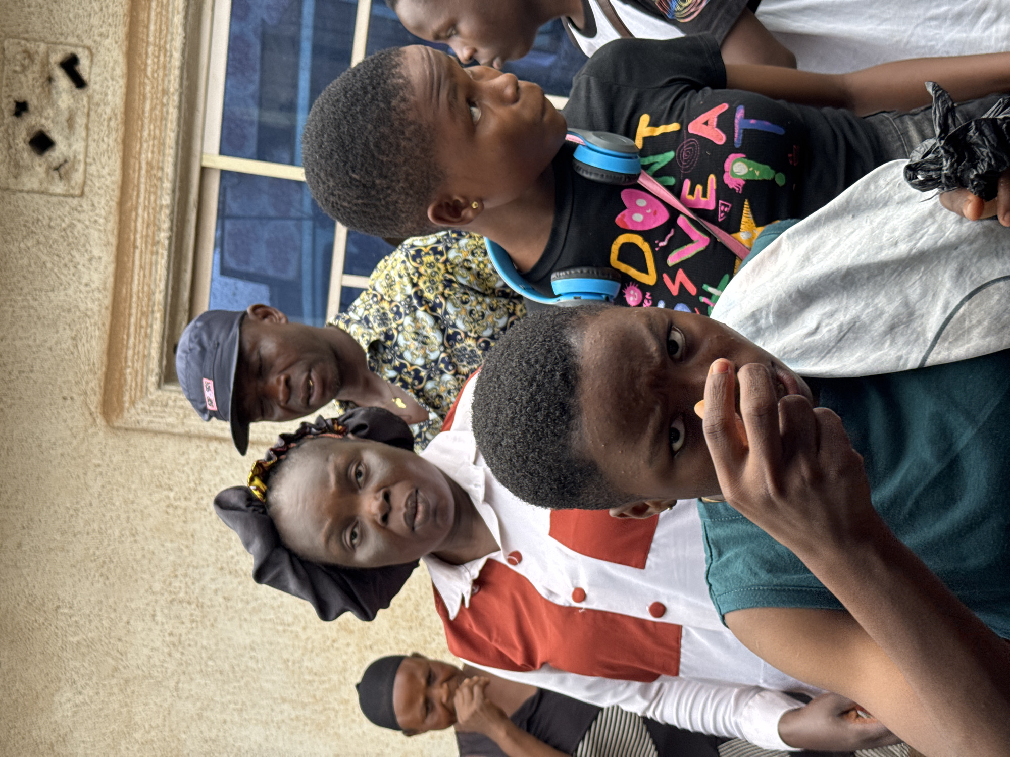 This image captures a group of people of varying ages, including children and adults, gathered in what appears to be an indoor or sheltered outdoor setting. The background features a textured, light brown wall and a window with blue-tinted glass panes.
In the immediate foreground on the right, a young person with short, dark curly hair looks directly at the camera with a curious expression. They are wearing a dark green t-shirt, and their left hand is raised near their mouth, holding a small, orange or yellow object.
Towards the center, a woman with dark skin wears a white shirt accented with red panels and red buttons, along with a black head covering. She is looking slightly upwards and to the left. Adjacent to her and further back, a man wears a blue baseball cap backward and a patterned yellow and black shirt, looking downwards.
Another young person is prominent in the mid-ground, wearing a black t-shirt with colorful, vibrant text that reads "DON'T SWEAT IT" alongside various symbols like hearts and stars in shades of pink, green, yellow, and blue. Blue headphones are resting around their neck, and a small gold earring is visible in their left ear. This individual is looking upwards and to the right.
In the background, partial views of other individuals are visible, including one young person in a white sleeveless top and another wearing a black head covering and a black and white striped shirt.
The scene is brightly lit, indicating it is daytime. The individuals' expressions vary, suggesting a relaxed communal gathering or waiting. The location context for this image is Oboama, Nigeria.