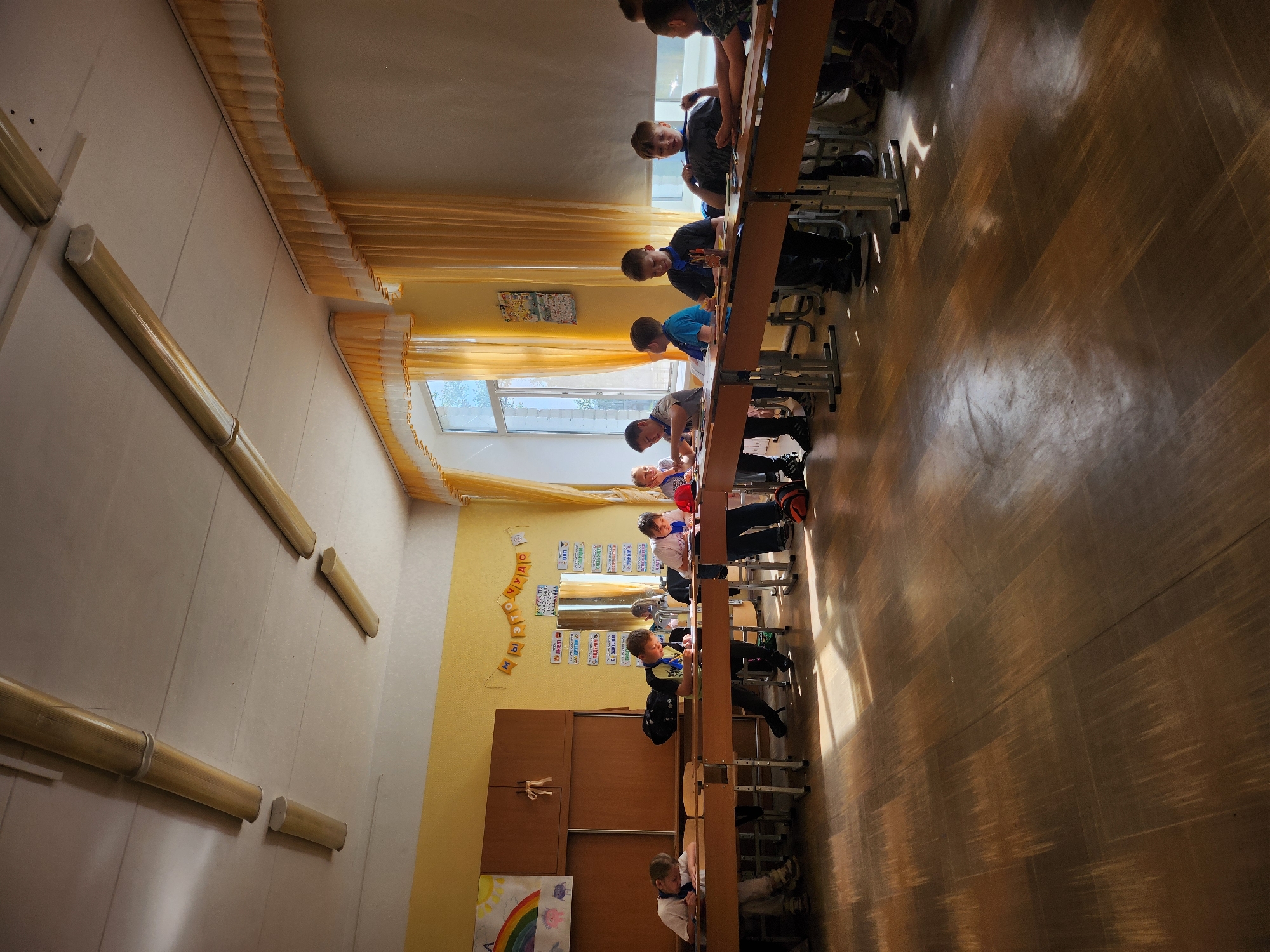 The image depicts a brightly lit classroom in Chaikovsky, Russia, during daytime, likely morning or early afternoon given the strong natural light and shadows. Approximately ten to twelve children, mostly boys of elementary school age, are present.
The classroom features rows of light-colored, adjustable student desks and chairs. Students are seated at the desks, appearing engaged in an activity, possibly listening, writing, or drawing. Some children are looking towards the front of the classroom (left side of the image when correctly oriented), while others are focused on their desks.
The room has a light-colored ceiling and walls, with one section of the wall painted a vibrant yellow. Large windows run along one side, covered with yellow or orange pleated curtains or blinds, allowing ample sunlight to stream into the room. This light casts distinct, long shadows across the wooden or wood-patterned floor.
Along the yellow wall, there's a brown wooden cabinet or set of cupboards. Above this, various educational posters and decorations are visible, including a child's drawing featuring a rainbow. A string of individual Cyrillic letters is hung as a banner, spelling out "МЫ" (We) and "ЧУДО" (Miracle/Wonder), with other unclear letters in between. Other posters with Cyrillic text are also visible but too blurry to decipher. The general atmosphere is calm and focused, typical of a school learning environment.
