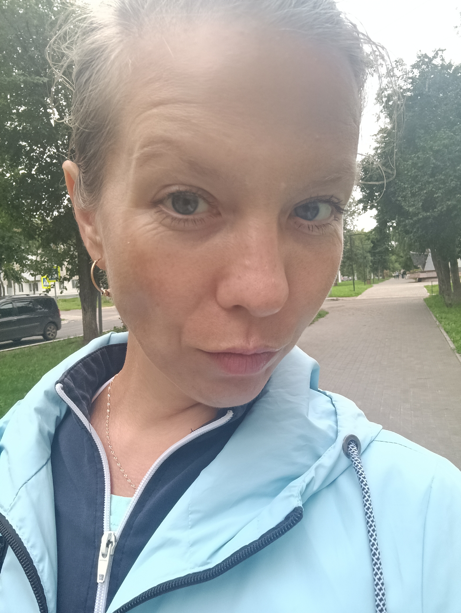 A woman, with light-colored hair pulled back and light-colored eyes, takes a selfie outdoors. She is wearing a light blue zippered jacket over a dark blue top. A thin gold chain necklace is visible. She has a hoop earring in her left ear. The background shows a paved walkway lined with trees and grass. A dark-colored car is parked on the left side of the image. The scene appears to be a public park or street in Chaikovsky, Russia, based on the style of buildings and street signage visible in the distance.