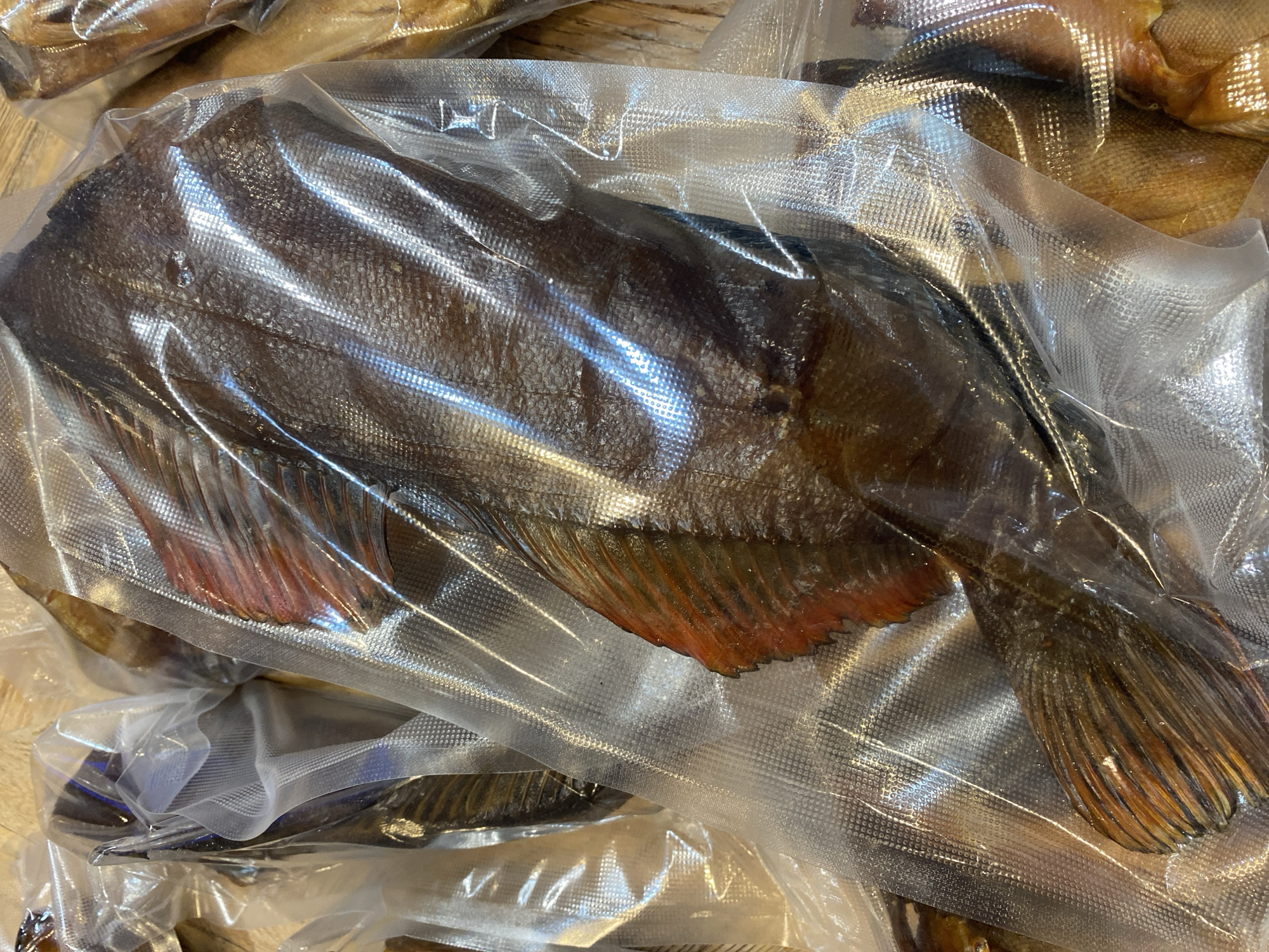 The image shows a close-up, overhead view of several smoked fish, vacuum-sealed in clear plastic packaging. The fish are arranged in a way that suggests they are being displayed or sold. The central fish is a large, dark brown specimen with visible scales and reddish-orange fins. Other fish, also in plastic packaging, are partially visible to the sides and in the background. The packaging itself has a textured pattern. The lighting creates reflections on the plastic. The setting appears to be a market stall or a retail display, with a wooden surface visible beneath the packaging. The location is Tret'ya Pad', Russia.