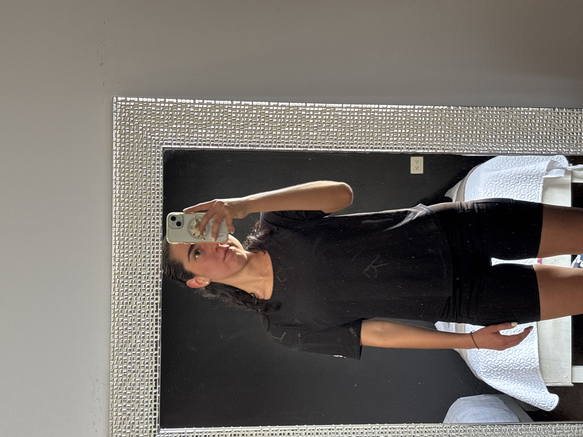 A young woman is taking a selfie in a mirror. The mirror has a silver, mosaic-tiled frame. The woman is wearing a black t-shirt and black shorts. She is holding a phone in her left hand. The background behind the mirror appears to be a wall and part of a room with furniture. The location is not discernible from the image.