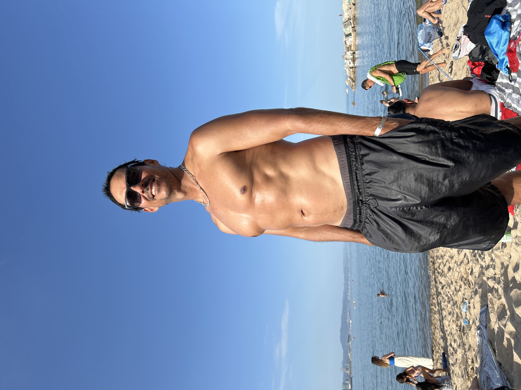 The image depicts a sunny day at a beach in Barcelona, Spain. The primary subject is a shirtless man wearing black swim shorts and sunglasses, standing on the sand and looking at the camera with a smile. He has a silver chain around his neck and his hands are in his pockets. In the background, the sea is visible with several boats and a distant cityscape. Other beachgoers are scattered across the sand and in the water, some swimming and others relaxing. The environment is characterized by clear blue skies, sand, and calm waters, suggesting a typical beach scene.