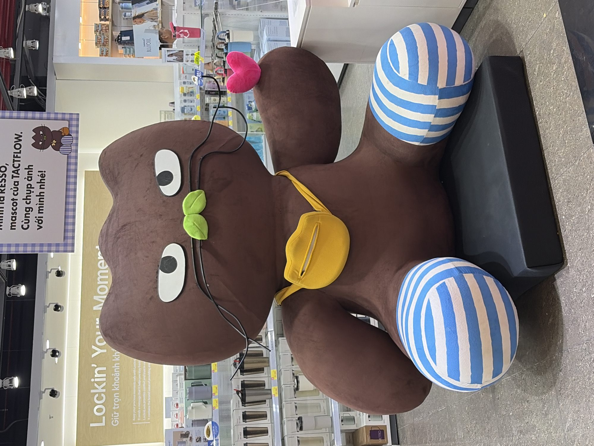 This image captures a large, plush, dark brown, stylized cat-like mascot situated in a retail store within Thủ Đức, Vietnam. The mascot sits prominently on a rectangular black platform on a grey tiled floor. It features large, white, oval eyes with black pupils, a small light green two-leaf-shaped nose, and thin black wires extending from its face, resembling whiskers or glasses. A bright yellow fanny pack or crossbody bag with a visible zipper is slung over its right shoulder. Its lower limbs are depicted as bulbous forms adorned with alternating horizontal blue and white stripes. The mascot holds a small, fluffy, bright pink heart in its left paw.
In the background, to the left, a promotional sign with a light yellow background displays "Lockin' Your Moment" in English, with "Giữ trọn khoảnh khắc" in Vietnamese beneath it, associated with the LocknLock brand. Below this sign, shelves are stocked with various insulated drinkware, including thermoses and cups in colors like silver, black, white, and green, with price tags displaying amounts in Vietnamese Đồng (e.g., "Đ 1.11.000"). To the right, a white and light blue checkered sign hangs, bearing Vietnamese text: "Minh là RESSO, mascot của TACTFLOW. Cùng chụp ảnh với mình nhé!" (Translation: "I am RESSO, TACTFLOW's mascot. Take a picture with me!"). This sign also includes a small cartoon illustration of a dark brown cat wearing striped pants, similar to the mascot. Further right, more retail shelving displays various products, and a partial sign for "BLI SOUL" is visible, along with a pink water bottle on a separate display. The ceiling features exposed track lighting with multiple white fixtures illuminating the scene. No human figures are present.