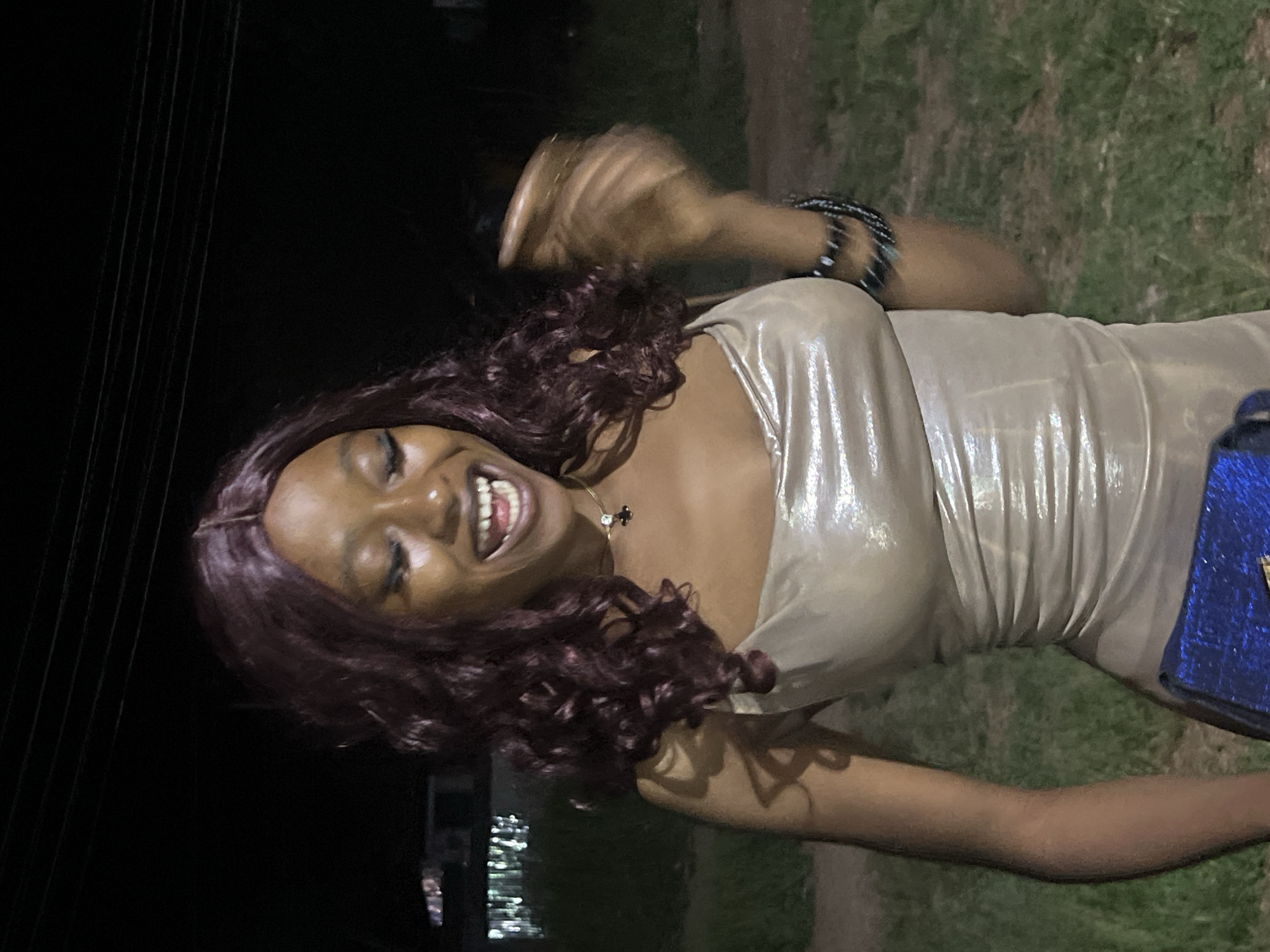 A young Black woman with auburn curly hair, a metallic champagne-colored slip dress, and dark makeup is captured in a medium shot outdoors at night. She is laughing with her mouth open and eyes closed, her right hand is blurred and extended forward, suggesting movement. She is wearing bracelets on her right wrist and a cross pendant necklace. A portion of a blue clutch is visible at the bottom of the frame. The background consists of dark grass and foliage, with faint lights from buildings and power lines in the distance, indicating an urban or suburban setting. The image appears to be taken in Durumi, Nigeria.