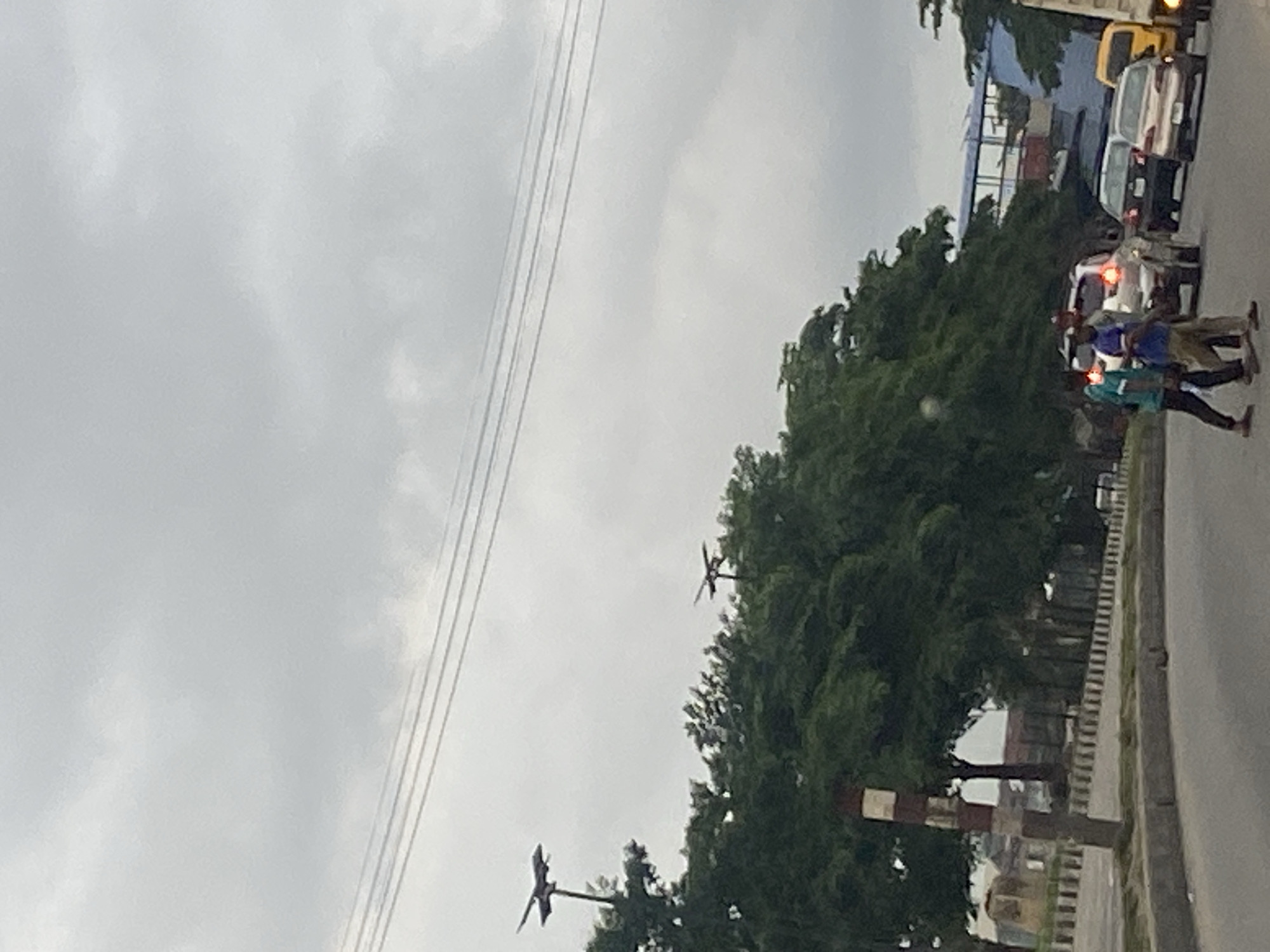 In Ikeja, Nigeria, on a cloudy day, the image captures a street scene with several vehicles and pedestrians. In the foreground, two young men are walking across a road, one in a turquoise shirt and the other in a blue and white striped shirt. Behind them, several cars are stopped or moving slowly in traffic, including a white SUV, a dark sedan, a light-colored sedan, and a yellow truck. Dense green trees line the side of the road, obscuring the view of buildings behind them. Overhead, power lines stretch across the sky. On the left side of the image, a striped pole stands near the road. In the distance, on top of the trees, two structures with fan-like shapes are visible, possibly solar-powered lights or decorations. The overall atmosphere is that of a busy urban environment experiencing overcast weather.
