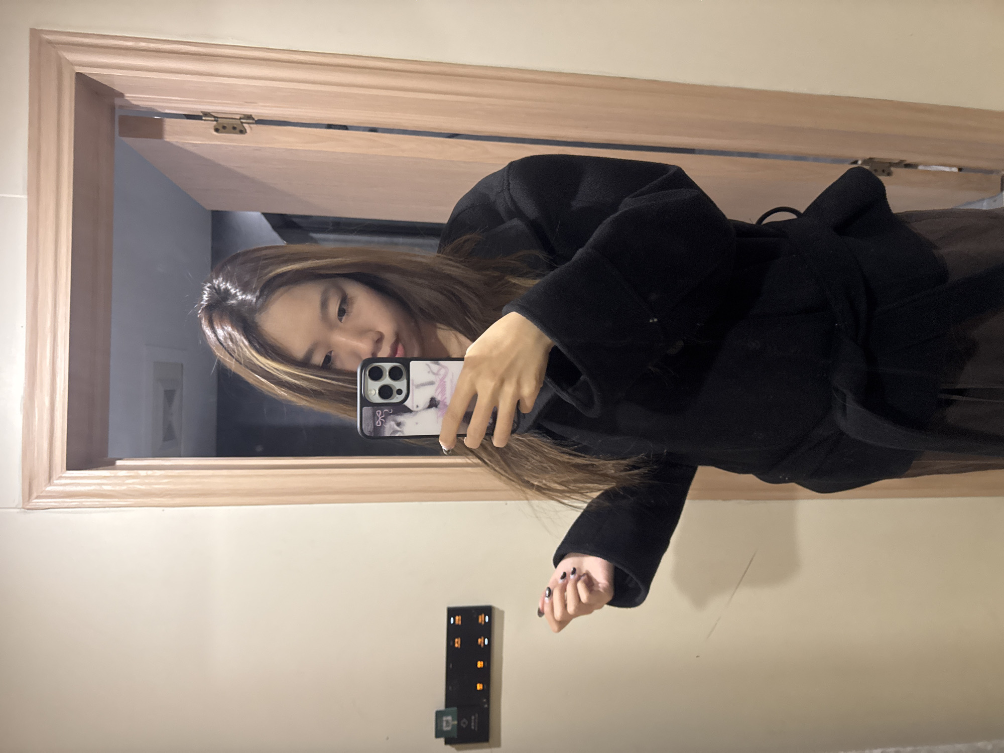 A woman is taking a selfie in a mirror. She is wearing a black coat and holding a phone with a patterned case. The mirror is framed with light-colored wood. In the reflection, a room with white walls can be seen. A black control panel with orange lights is visible on the wall next to the mirror. It is impossible to determine the location as Dunhuang City, China from the image.