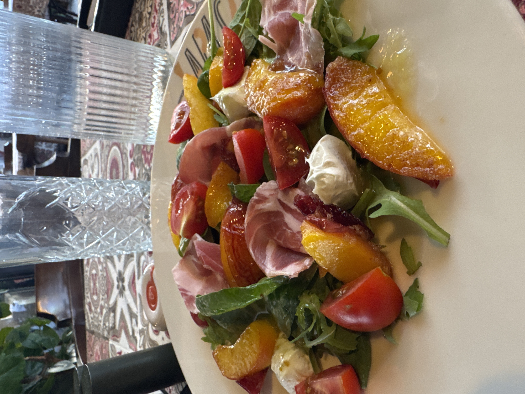A close-up, overhead shot shows a salad on a white plate. The salad contains sliced peaches, cherry tomatoes, prosciutto, and mozzarella cheese, all resting on a bed of arugula. The plate is on a patterned tablecloth. In the background, two clear glasses of water, one with a diamond-cut pattern and the other with vertical ridges, are visible. A plant and part of a chair can be seen further back, suggesting a restaurant setting. The lighting appears to be natural, coming from a window or overhead source. The salad is the primary focus of the image, with the surrounding elements slightly out of focus. The image was taken in Chernivtsi, Ukraine.