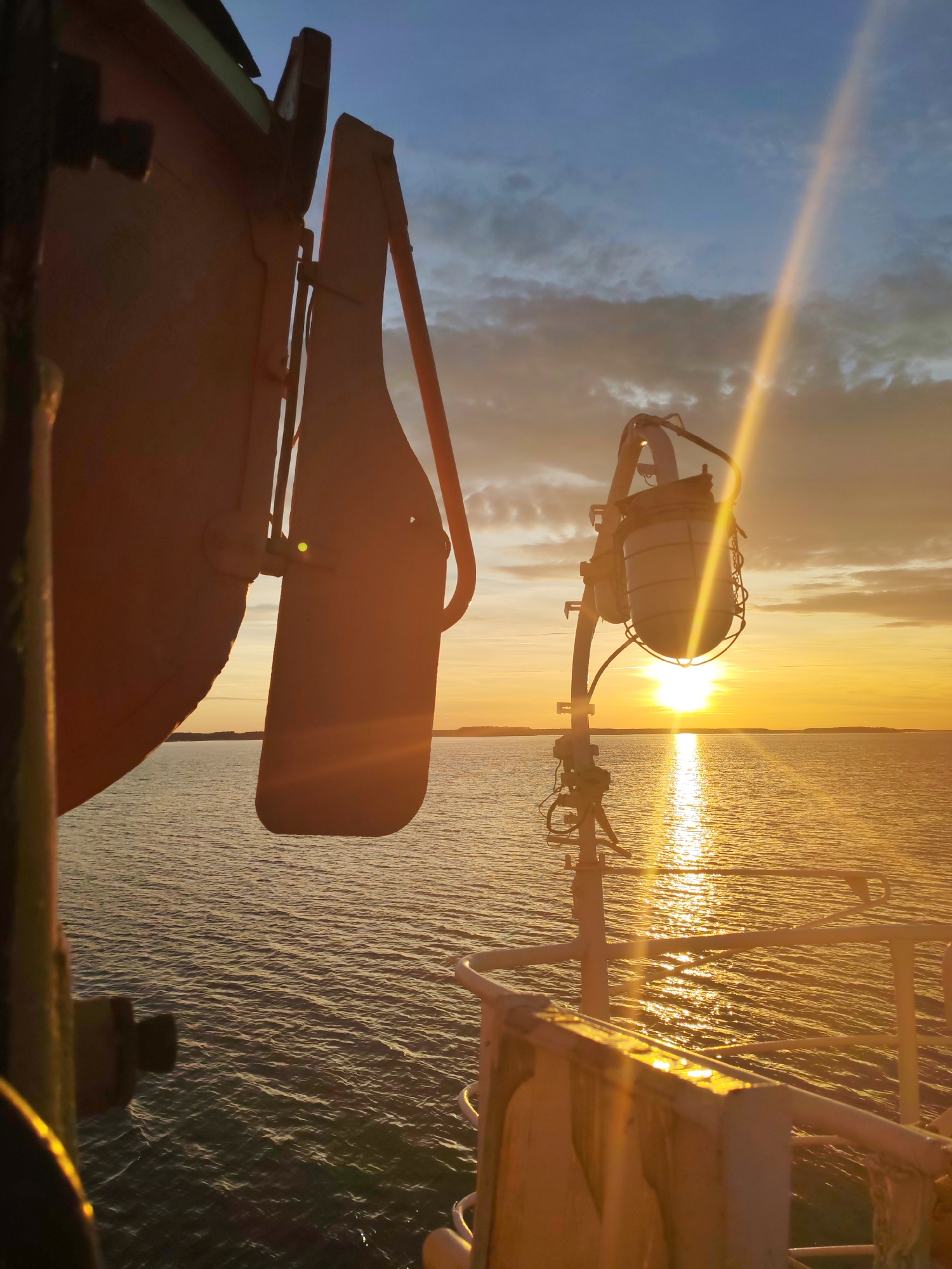 The image depicts a sunset over a body of water, possibly a sea or large lake, from the perspective of a vessel. The sun is low on the horizon, casting a golden light across the water. To the left, a lifeboat or similar emergency craft is visible, mounted on a davit. A lamp post with a protective cage stands to the right, and a portion of the vessel's railing can be seen. The location is unknown. No people are visible. The scene evokes a sense of tranquility.