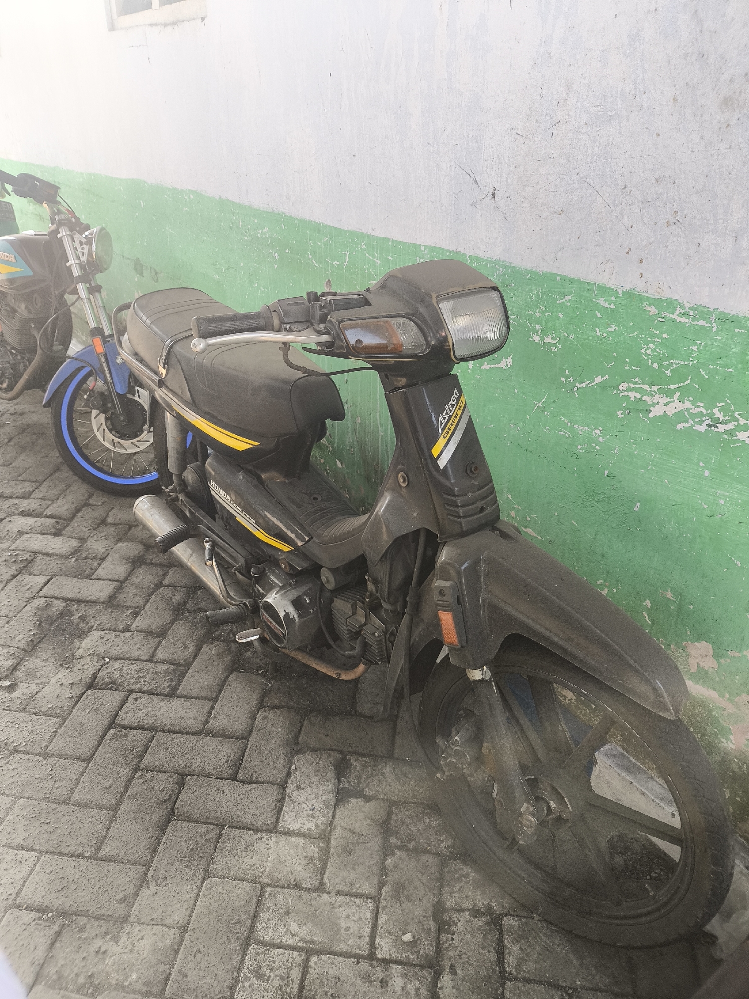 Two motorcycles are parked on a paved surface in front of a wall. The wall is painted in two horizontal sections, a lighter color on top and a faded green on the bottom. The motorcycle in the foreground is black with yellow accents and appears to be a Honda Astrea Grand, covered in a layer of dust. The second motorcycle, behind and to the left, is blue and silver, with a blue stripe around the rim of its front wheel. Both motorcycles are stationary. The scene appears to be an outdoor environment, possibly a residential or public area in Lumajang, Indonesia. The lighting suggests it is daytime.