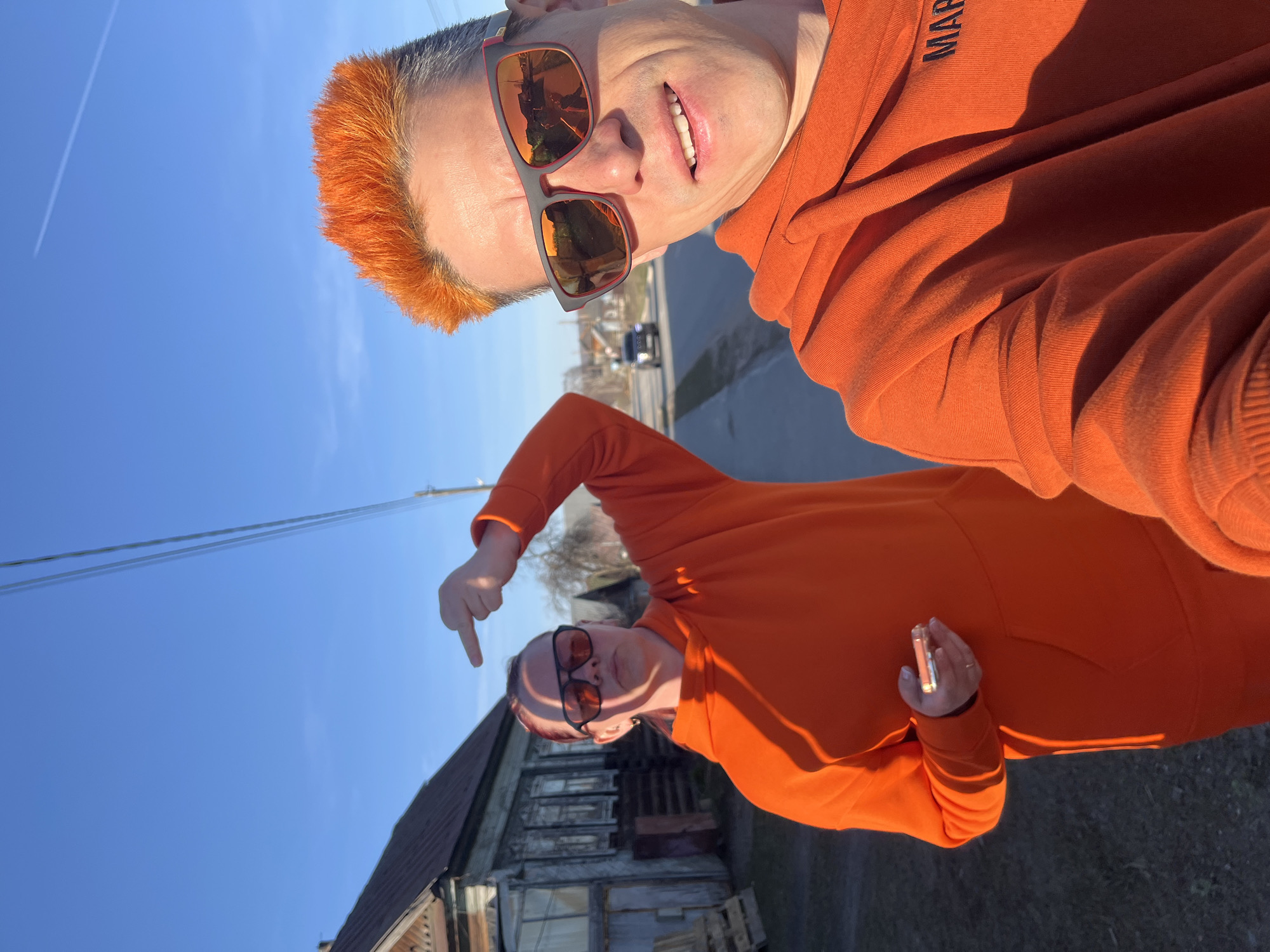 The image features two instances of a person, likely the same individual duplicated or reflected, set against an outdoor, semi-urban background under a clear, bright blue sky in Chaikovsky, Russia.
The upper figure, oriented vertically, appears to be taking a selfie. This person has short, spiky bright orange hair and is wearing sunglasses with orange-tinted lenses and dark frames, reflecting the surrounding environment including a street and a distant vehicle. A slight smile is visible, and they are dressed in an orange hooded sweatshirt with dark text, possibly "MAP" or "MARP," visible on the shoulder. A white contrail streaks across the upper left portion of the blue sky.
The lower figure, angled diagonally, also wears an identical orange hooded sweatshirt and dark sunglasses. This individual is pointing downwards with their right hand. In their left hand, they hold a light-colored smartphone. They are positioned in front of an old, rustic-looking building with a dark roof and weathered wooden walls, featuring several windows that appear to be covered or boarded. Overhead power lines run across the sky above the building. The strong shadows indicate bright daylight and sunny weather.
Both figures showcase a casual style, with the vibrant orange attire standing out against the muted tones of the old building and the bright sky.