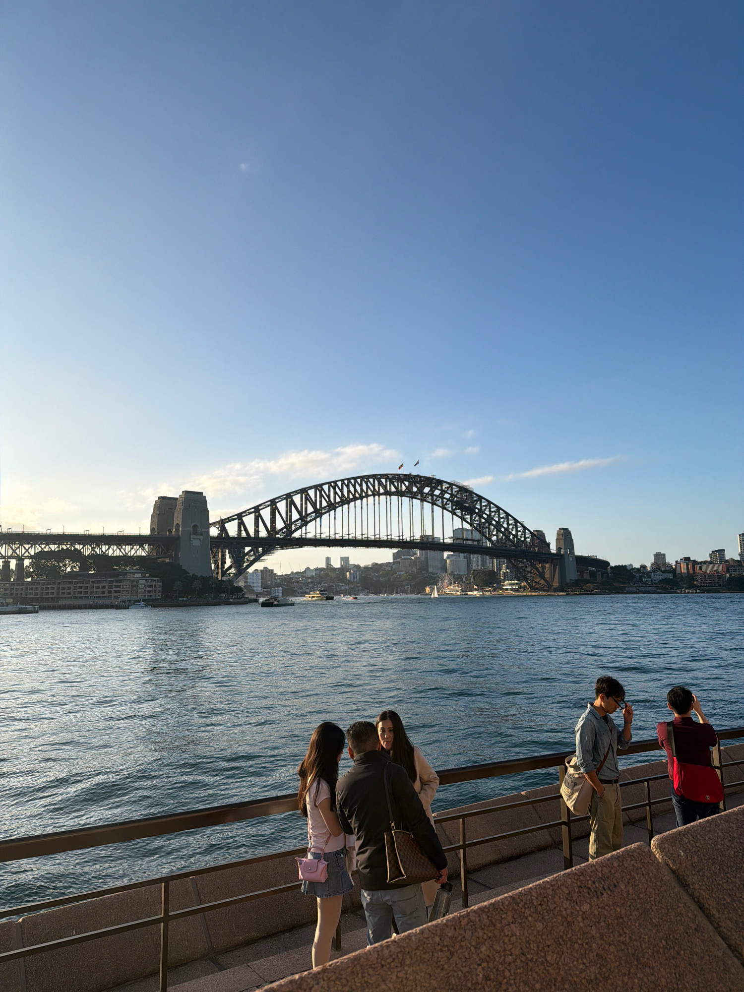 The image depicts a view of Sydney Harbour Bridge in Sydney, Australia, under a clear blue sky with a few wispy clouds. The sun is casting a warm light on the scene, illuminating the water of the harbour and the iconic steel arch bridge. Several people are visible on a walkway in the foreground, observing the bridge and the surrounding cityscape. On the left, a couple stands close together, with the woman wearing a pink shirt and denim skirt, and the man in a dark jacket and jeans. To their right, a man in a light blue shirt and khaki pants is looking down, adjusting his sunglasses. Further to the right, another man in a maroon shirt and dark pants is taking a photo with his phone. In the background, various buildings of the Sydney skyline are visible, along with boats on the water, including a ferry. The water is calm, with gentle ripples reflecting the light. The walkway has a metal railing and a stone ledge. The overall atmosphere is one of a pleasant day in a vibrant urban environment.