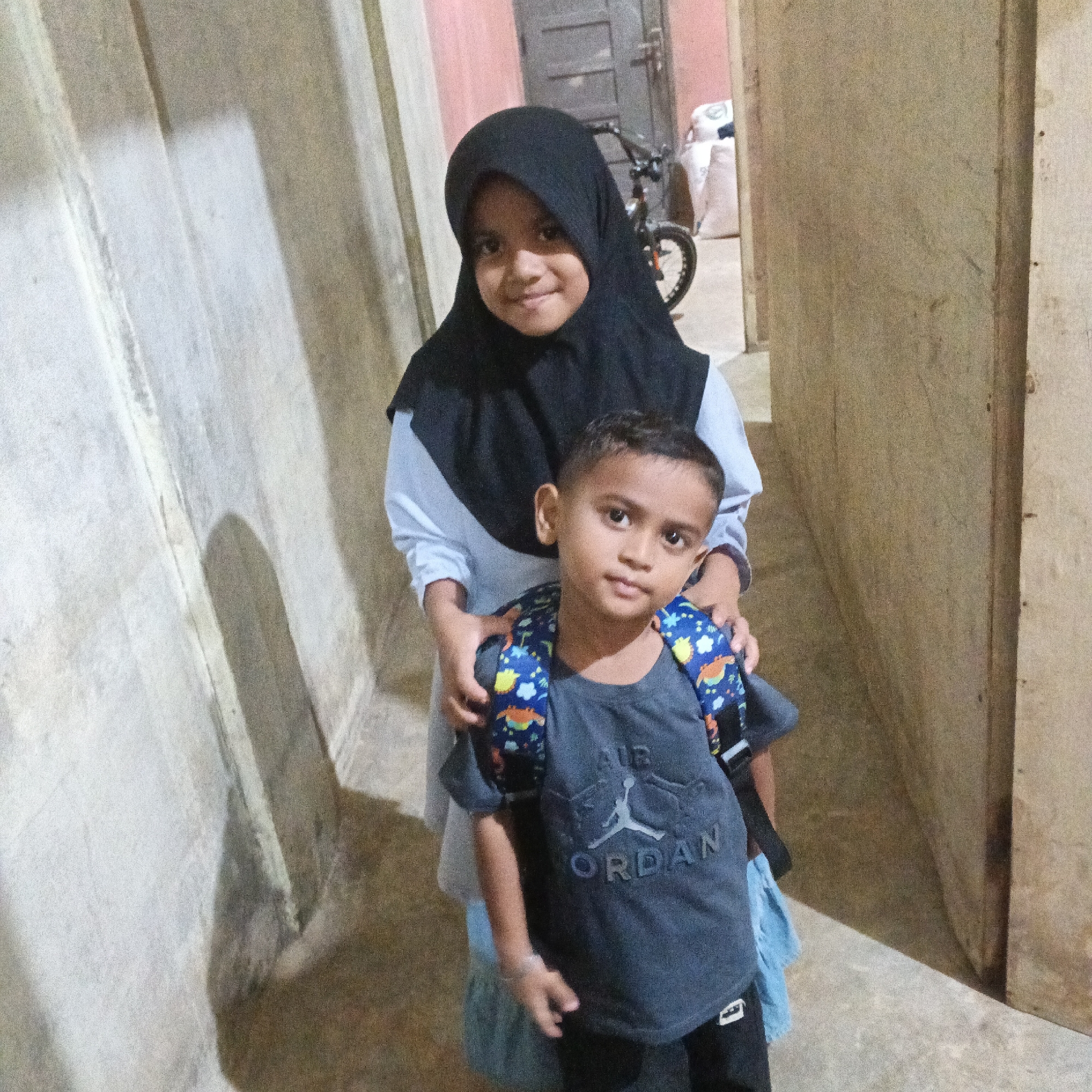 Two children, a girl and a boy, stand in an indoor hallway. The girl, on the left, wears a black hijab and a light blue long-sleeved shirt. Her hands are resting on the boy's shoulders. The boy, in front, wears a dark grey t-shirt with a white "Jordan" logo and a backpack with a colorful pattern. He is looking directly at the camera with a slight smile.
The setting appears to be a simple, rustic interior, possibly a hallway or passageway. The walls are textured, with one side appearing to be made of rough, pale material and the other of rough, darker wood. A shadow is cast on the pale wall, suggesting a light source from the right. In the background, a bicycle and some large white bags are visible, hinting at a storage area or a functional space. The lighting is dim, suggesting it could be daytime indoors, or possibly evening. The overall atmosphere is casual and domestic. There is no indication of the weather. The children's expressions are calm and content.
The visible text on the boy's shirt reads "JORDAN". The location context is Meunasah Bie, Indonesia.