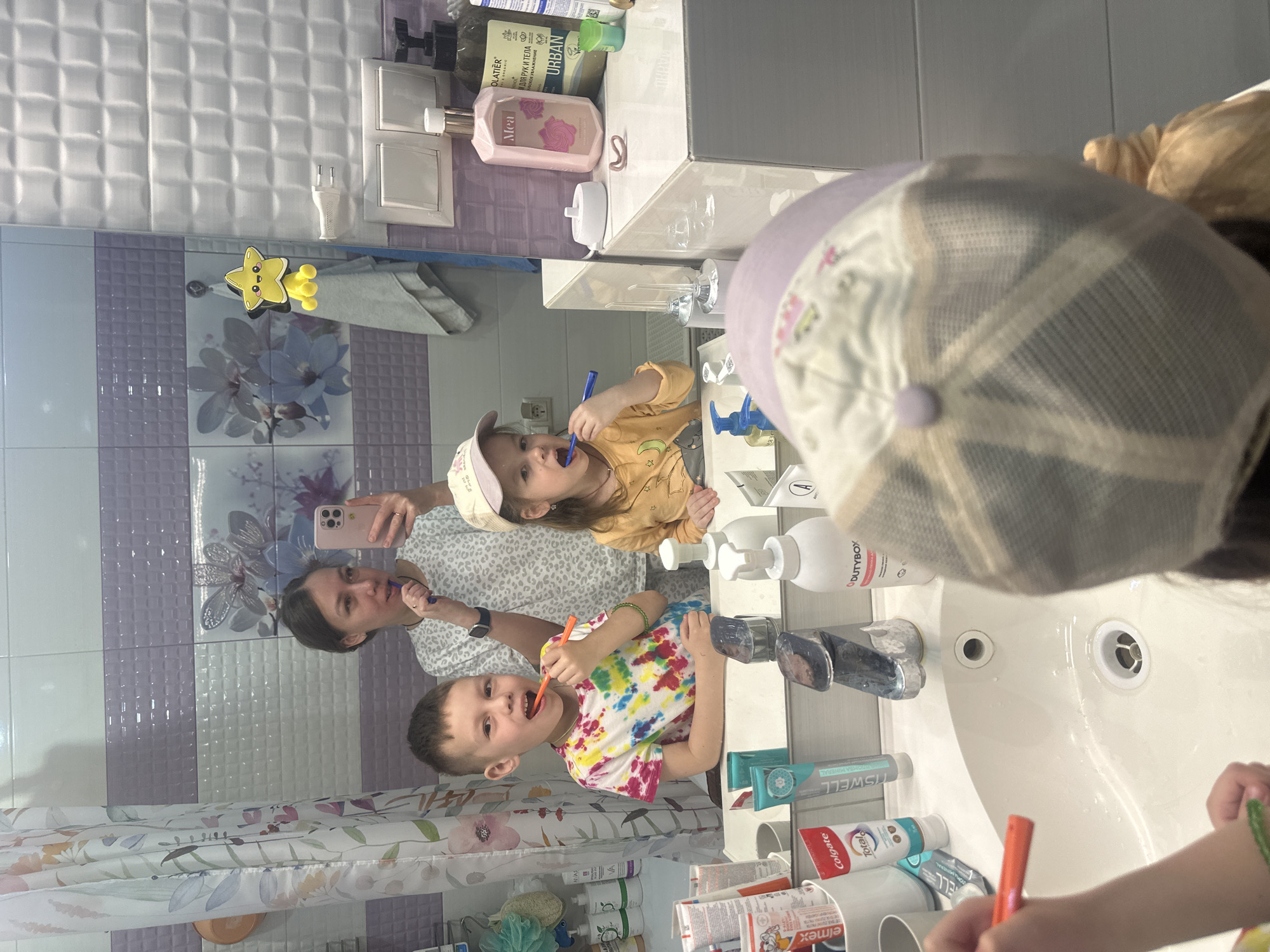 A mirror selfie is being taken in a bathroom. A woman is holding a cell phone, brushing her teeth in the mirror along with her children. The children, a boy and a girl, are also brushing their teeth, with toothbrushes of different colors. The bathroom has lavender and white tiles. Various toiletries and toothpastes are on the counter. A shower curtain hangs to the side. Location would be the city of Tomilino, Russia.