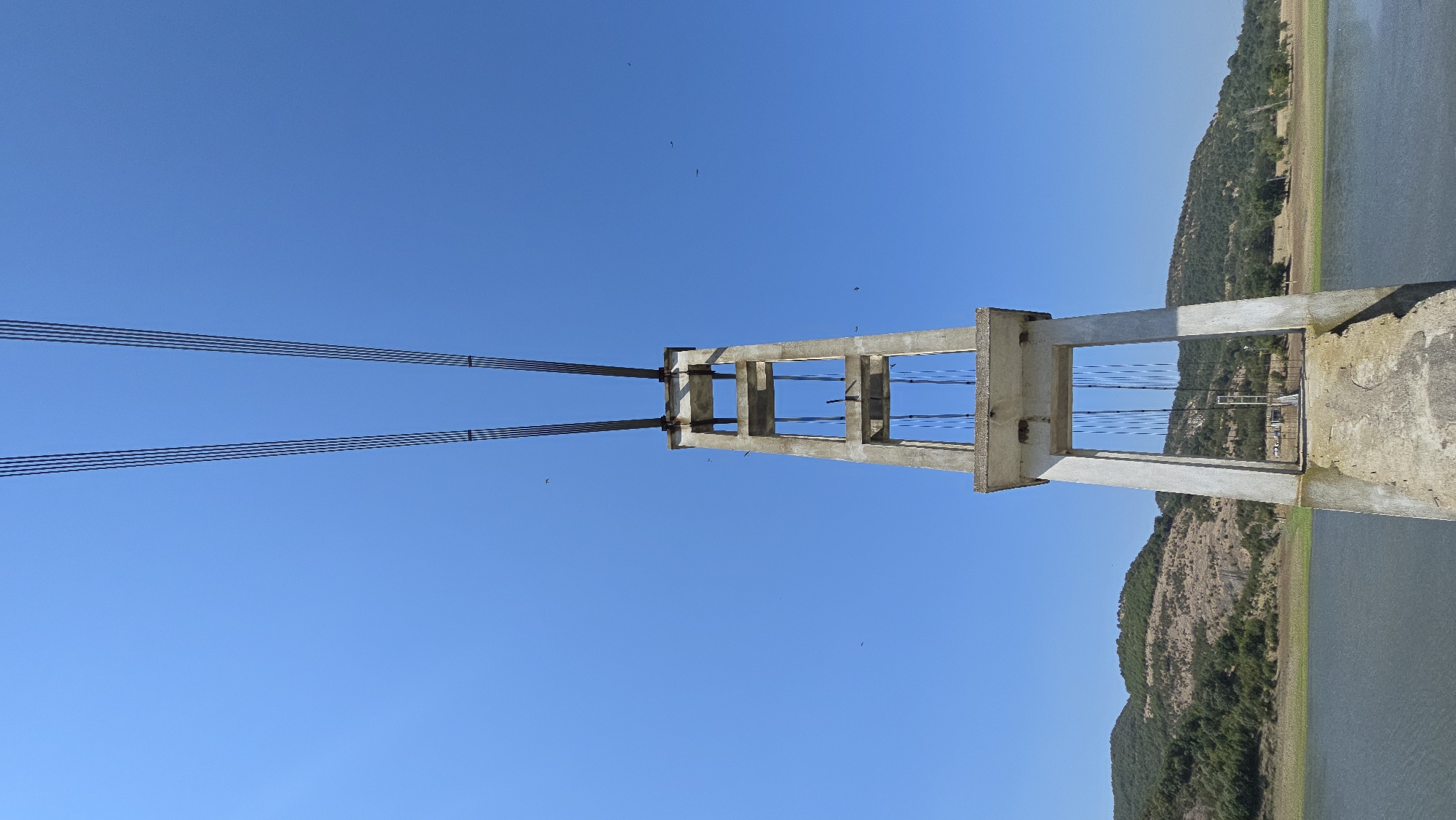 A concrete pylon stands tall against a clear blue sky, its weathered structure supporting thick steel cables that stretch upwards and away from the frame. The pylon features an open, rectangular design with multiple levels, and the cables are visible within its structure, descending towards the unseen ground. Below, a body of calm, dark water stretches to the horizon, meeting a gently sloping shoreline dotted with sparse vegetation. In the background, rolling hills covered in green trees and dry brush rise against the sky, their slopes showing signs of erosion in places. The scene is devoid of human presence, with only a few small, dark specks, likely birds, visible in the sky. This image captures a section of an abandoned or incomplete suspension bridge structure located in the vicinity of Lisitsite, Bulgaria.