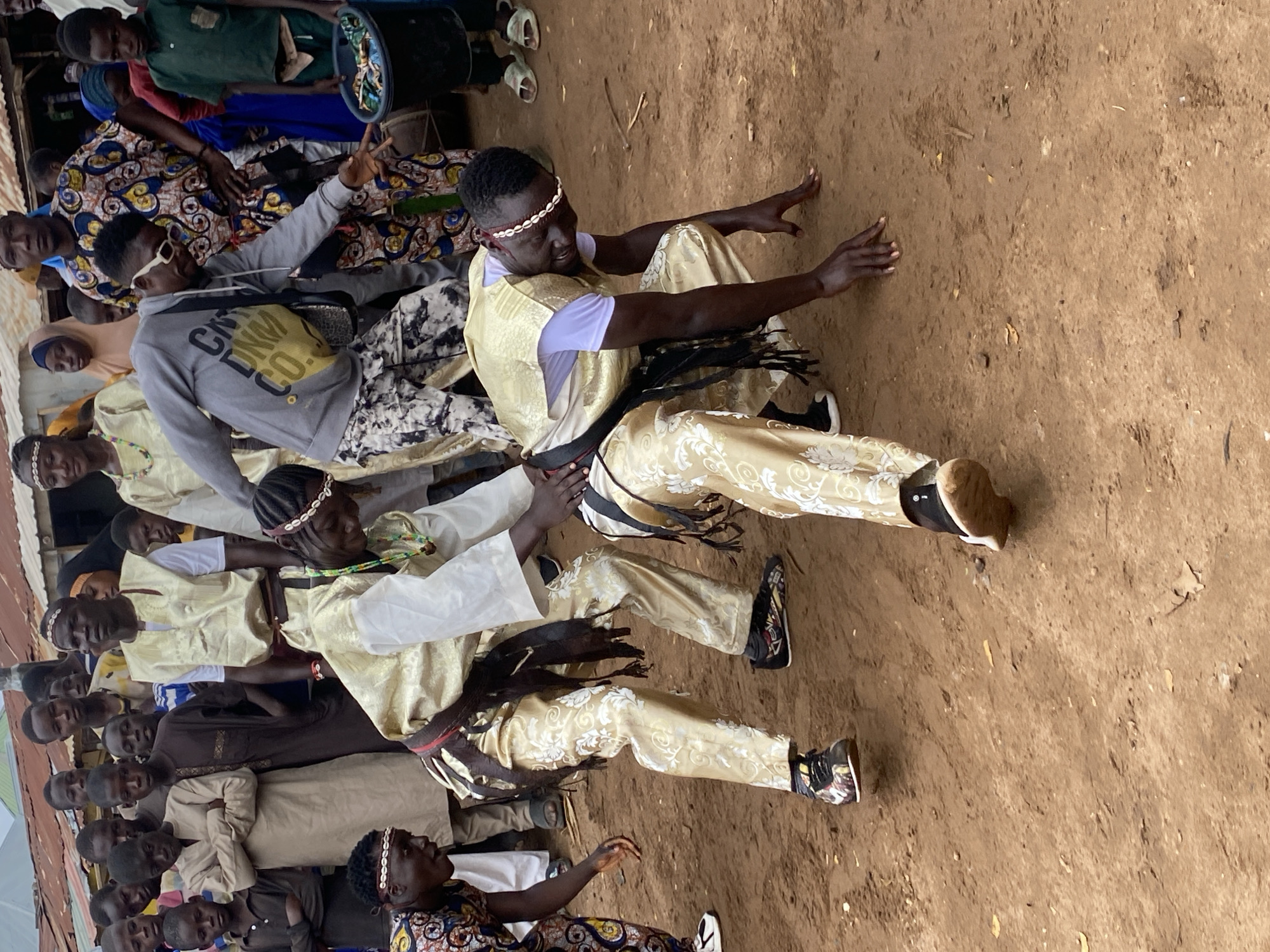 In Rigachikun, Nigeria, two young men in traditional gold attire are engaged in a dance. One performer is crouched low, hands on the ground, his face contorted in a dance move. The other, standing, assists him, one hand on the crouched performer's back, the other extended. Both wear beaded headbands and the standing performer has a dark sash around his waist. Other villagers, including children, watch the performance in the background, some wearing similar traditional clothing. The ground is dusty earth, with a few dry leaves scattered around. In the background, makeshift structures suggest a rural or semi-urban setting.