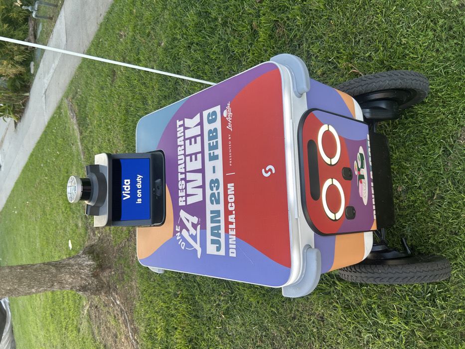 An autonomous delivery robot is positioned on a grassy lawn, promoting "DINE LA Restaurant Week" from January 23rd to February 6th. The robot features a screen displaying "Vida is on duty" and a vibrant design with the event's details. Presented by Los Angeles, the promotion emphasizes dinela.com and includes a stylized face-like graphic on its side. The robot's wheels are visible, highlighting its mobility.