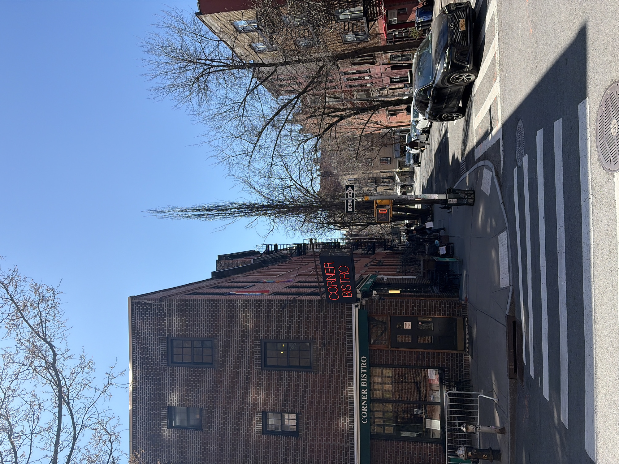 The image depicts an outdoor scene in New York City, United States. The "Corner Bistro" restaurant is the prominent subject, featuring a brick building with a green awning and neon signage. The establishment is situated at a street corner with a crosswalk marked with white stripes. Adjacent to the crosswalk is a parked dark-colored car. The sky is clear and blue. Deciduous trees with bare branches are visible on both sides of the street. Other buildings and cars are visible further down the street. There are street signs and a traffic signal present. People are also present at the restaurant and on the street.