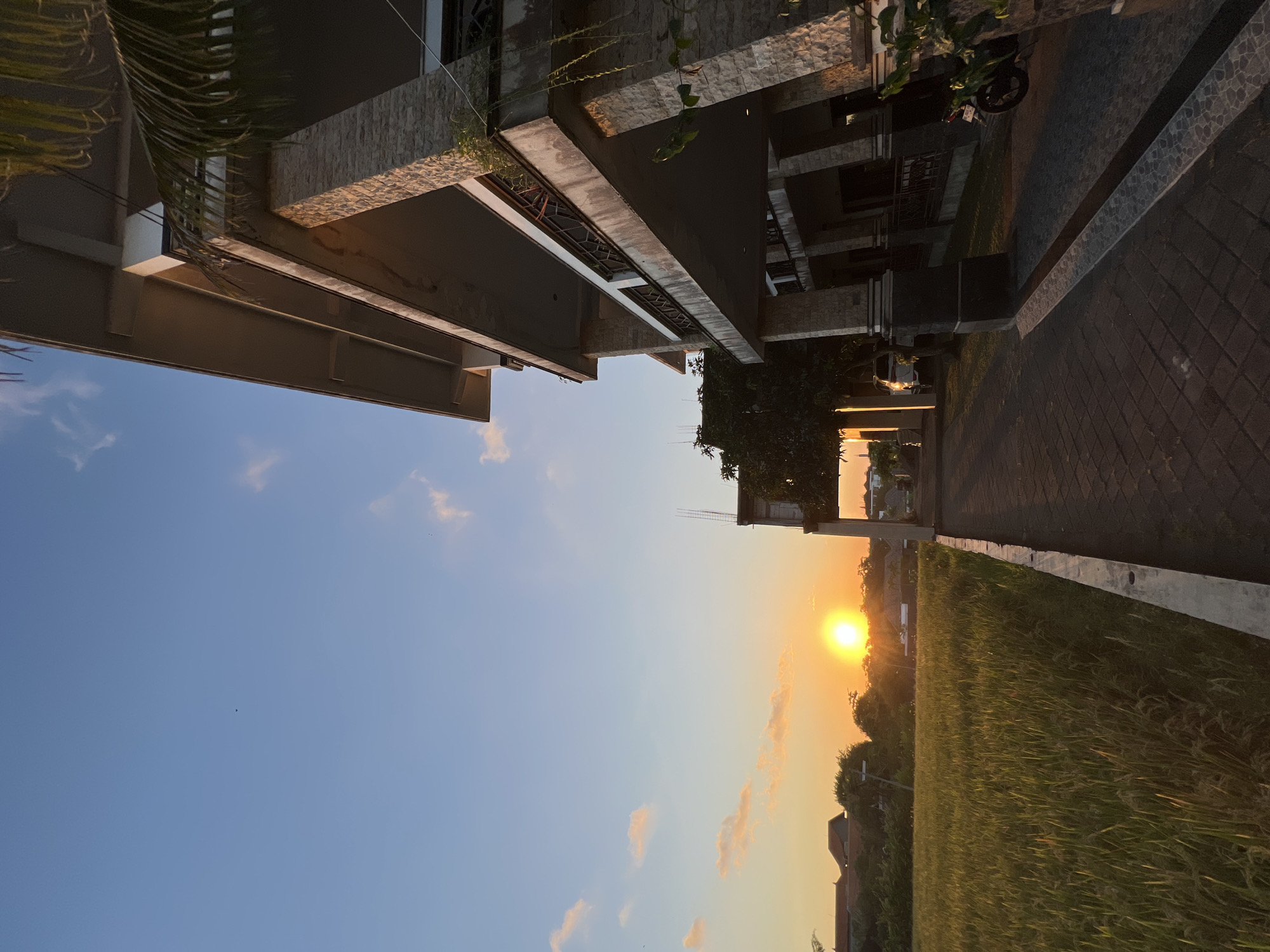 The image depicts a sunset scene in North Kuta, Indonesia. The sky is a gradient of blue to orange, with the sun setting on the horizon behind a field of tall grass. Two buildings frame the top of the image. The building on the left has a modern, angular design with visible windows and textured stone elements. The building on the right also has a similar style with a brick walkway. Part of a motorcycle and plants are visible near the building on the right. The scene suggests a residential area close to rice fields.