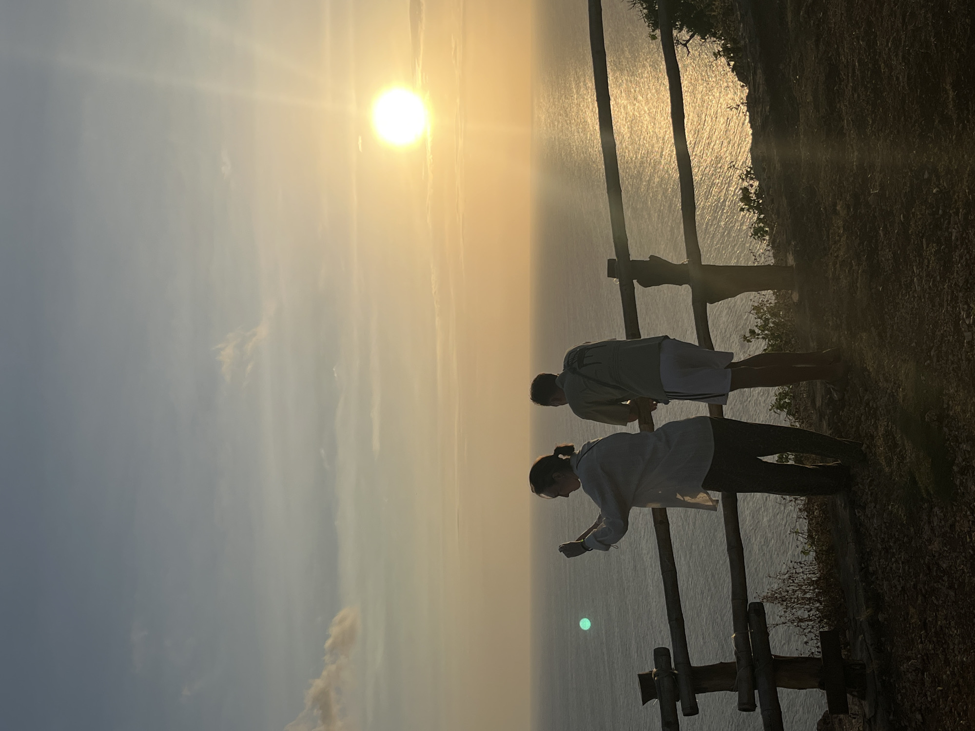 This is a vertically oriented, low-angle, outdoor photograph of two people standing on a cliff overlooking the ocean at sunset. The sun is bright and low in the sky, casting a warm, golden light across the scene. The water below reflects the sunlight, creating a shimmering effect. The figures of the two people are silhouetted against the bright sky, making their features indistinct. The person in the foreground is a woman wearing a light-colored top and dark pants, and she is holding a phone in her hand. The person behind her is a man wearing a light-colored top and shorts. They are both looking out at the ocean. There is a rustic wooden fence in the foreground, and on the right side of the frame, there is a rocky cliff face with some vegetation. The scene suggests a peaceful and contemplative moment, possibly at the end of a day of sightseeing or relaxation. The location is Kuta Selatan, Indonesia, which is known for its beautiful beaches and sunsets. The weather appears to be clear and calm. The time of day is sunset, indicated by the low sun and warm lighting. No text is visible in the image.