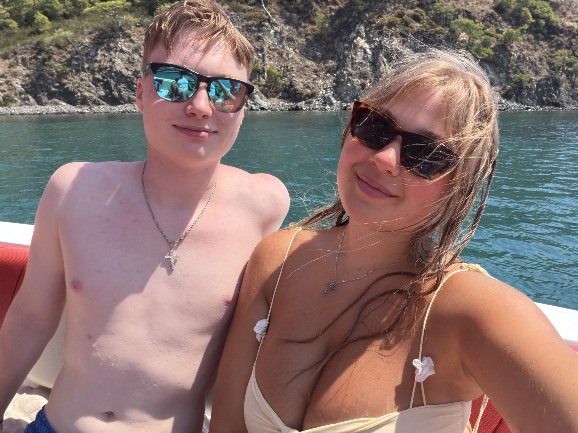 In Kemer, Turkey, a young Caucasian man and woman pose for a selfie on a boat. The man, on the left, wears blue swim trunks, a silver chain with a cross pendant, and mirrored sunglasses. The woman, on the right, has blonde hair, a tan, and wears a cream-colored bikini top with decorative floral accents. She also wears sunglasses and a silver chain with a cross pendant. The background features the turquoise waters of the Mediterranean Sea and a rocky, tree-covered hillside characteristic of the Kemer coastline. The scene is brightly lit by sunlight.