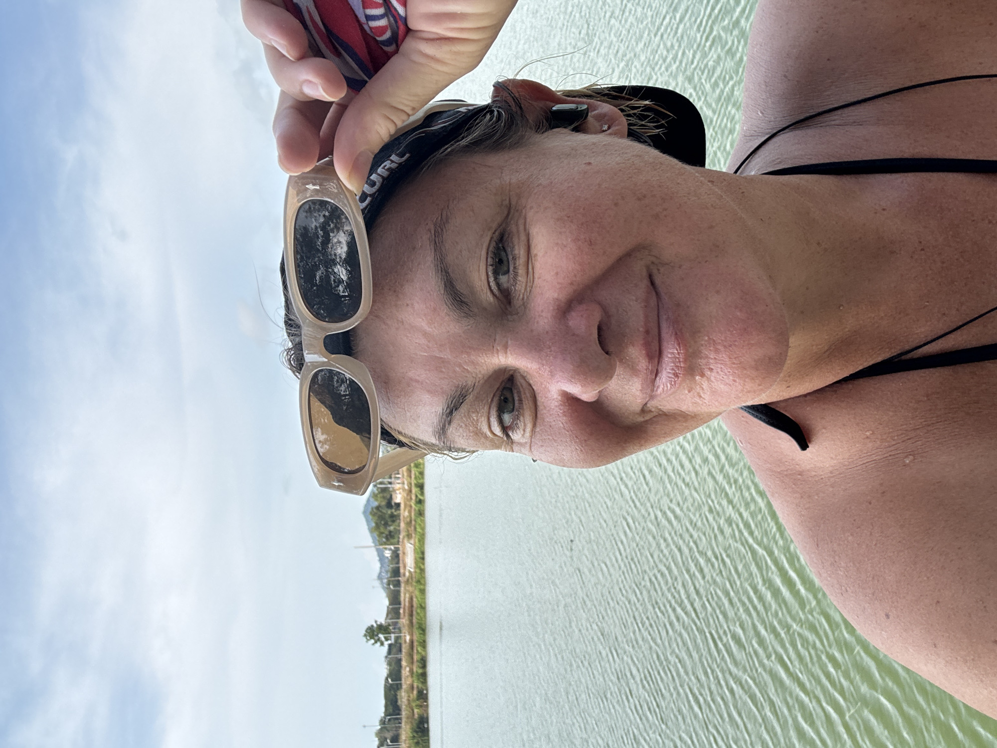 A selfie of a middle-aged woman with wet hair wearing a black swimsuit with her sunglasses in her hand. She is near the water, with vegetation and buildings in the background. The location is identified as Baan Chaweng, Thailand based on background details. The water reflects the cloudy sky. The woman is looking at the camera with a slight smile.