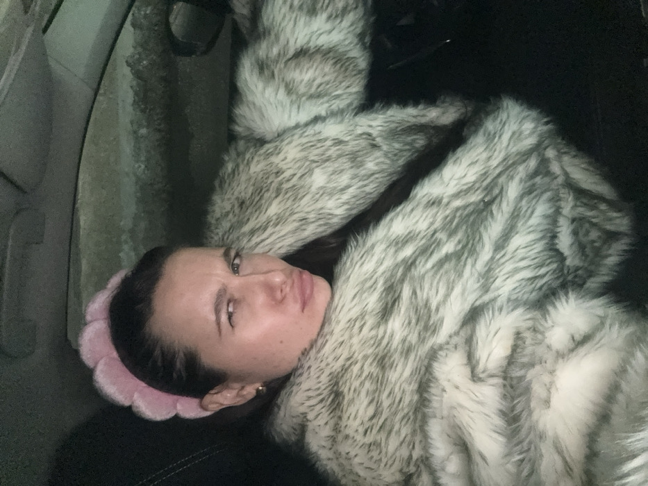 A single female individual is seated within a vehicle, observed in Akseno-Butyrki, Russia. She has dark hair styled back and wears a pink, scalloped headband. A small gold hoop earring is visible on her left ear. She is attired in a voluminous grey and white faux fur coat. Her facial expression features pursed lips and a slight squint of her right eye, suggesting a look of skepticism or mild displeasure. The vehicle's interior is dark, with a black seat visible. A layer of snow or ice is apparent on the exterior of the vehicle's side window, indicating cold ambient conditions. The scene is illuminated by dim, artificial light.