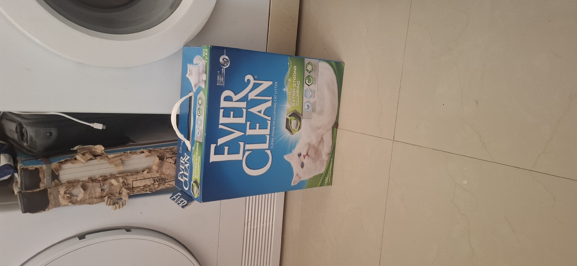 A box of Ever Clean brand cat litter is positioned on a tiled floor in front of two white appliances, likely a washing machine and a dryer. The cat litter box is predominantly blue with white lettering and features an image of a white cat. The box has a white handle on top. To the left of the cat litter box, a portion of a damaged cardboard box is visible, with torn edges and exposed corrugated material. Behind the cat litter box, a black object with a white cable is partially visible. The environment appears to be indoors, possibly a laundry room or utility area. The floor is made of light-colored square tiles. No people are present in the image. The scene is located in the city of Yerocham, Israel.