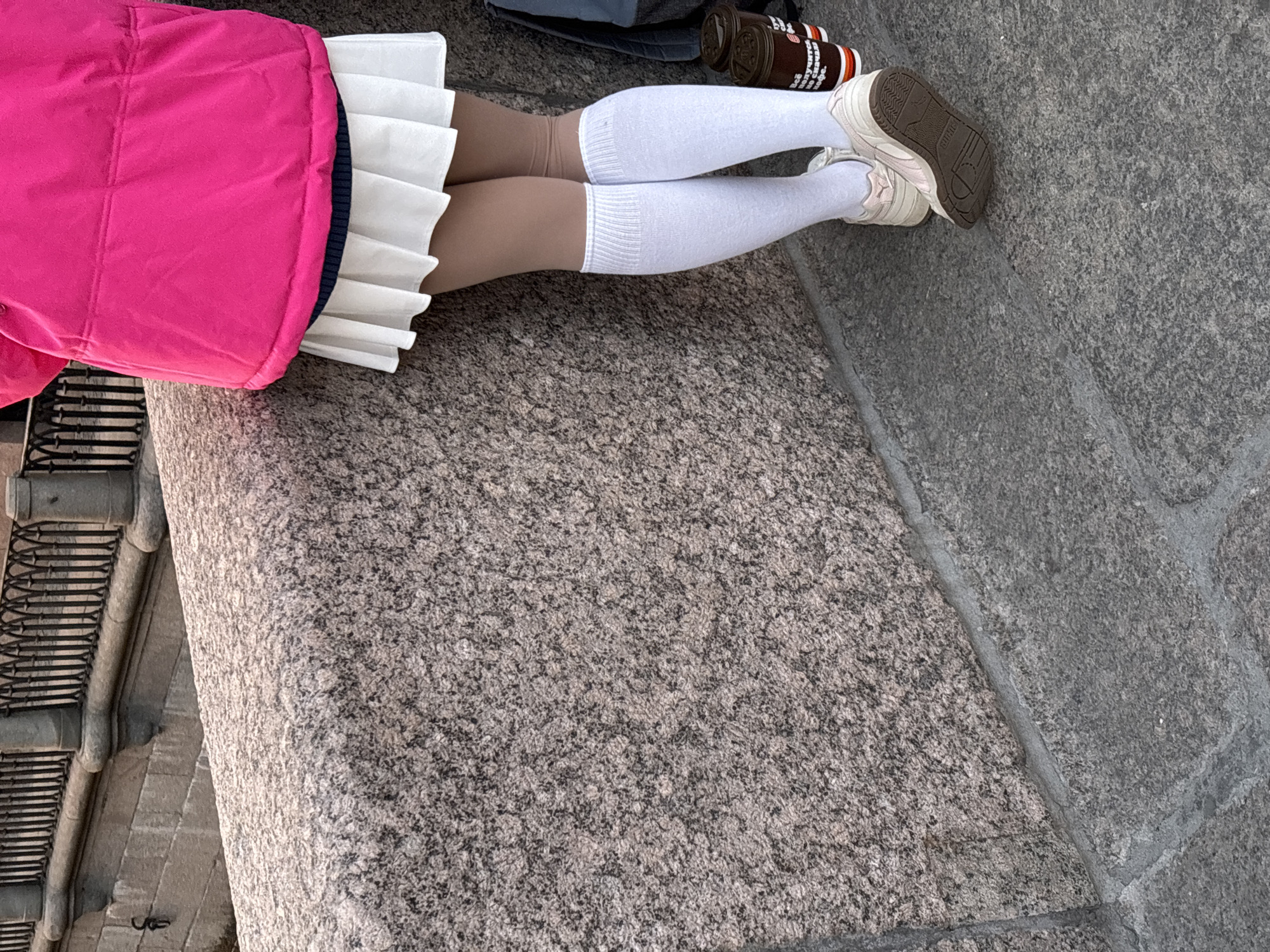 The image shows a person with a pink jacket, a white pleated skirt, white socks, and tan-colored skin on their legs, lying on a granite surface. Next to their leg are two cans or bottles. The granite surface appears to be part of a structure, possibly a step or ledge, with metal railings visible to the left. Based on the style of the architecture and materials, the location is suggestive of Saint Petersburg, Russia.
