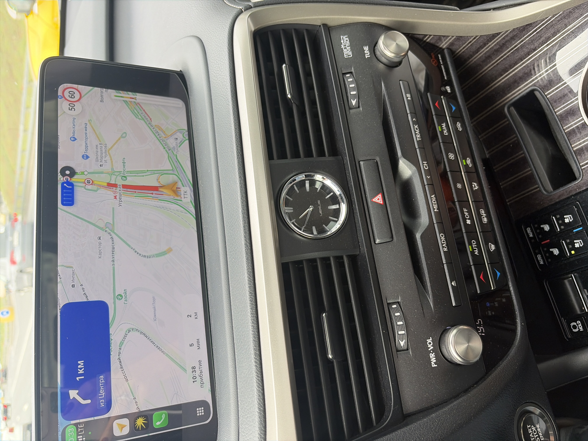 The image shows the interior of a Lexus vehicle, likely in Moscow, Russia, as indicated by the road signs and Cyrillic text on the navigation screen. The navigation screen displays a map with a route highlighted, indicating 1 km to the center and an estimated arrival time of 10:38, with 5 minutes and 2 km remaining. Speed limit indicators of 50 and 60 are visible on the screen. The center console features an analog clock with "LEXUS" branding, air vents, and various buttons and knobs for climate control and audio functions. A "PWR-VOL" knob and a temperature display of "19.5" are visible. Buttons for "RADIO," "MEDIA," and climate control modes like "AUTO," "OFF," "DUAL," and seat heating/cooling are also present. The "START STOP ENGINE" button is visible on the left side of the console. The overall scene captures a snapshot of a car's dashboard while in transit.