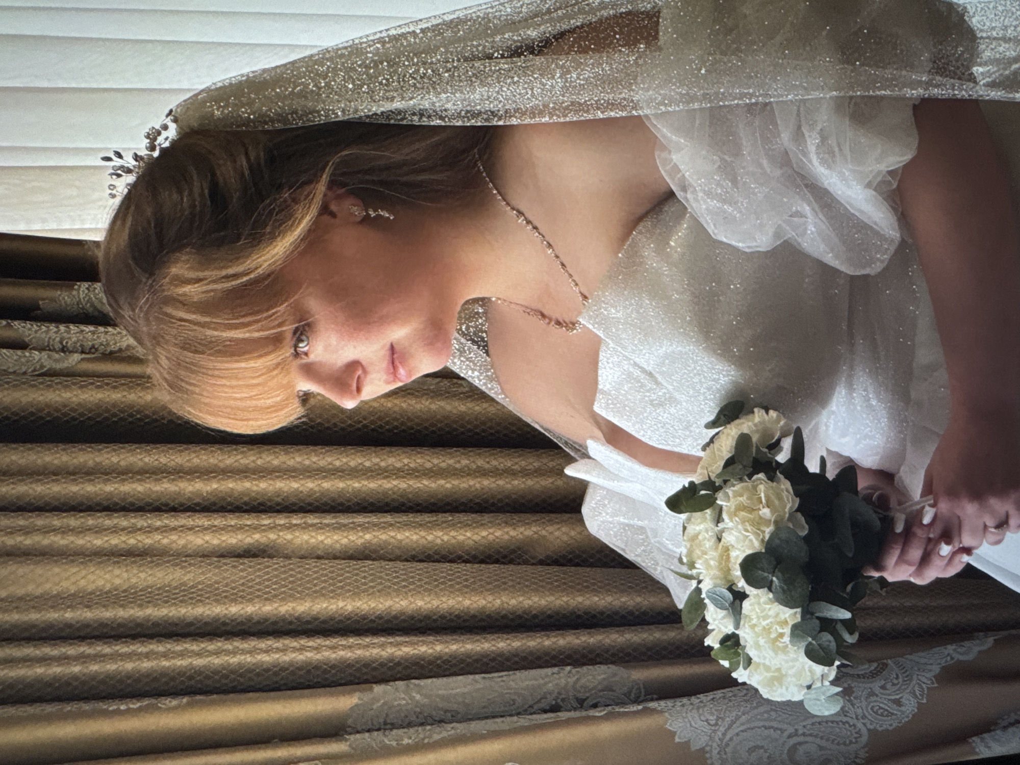 A woman in a wedding dress and veil is holding a bouquet of white flowers. She is wearing a necklace and earrings. The background is a curtain. The location is unknown, but it is supposedly near the city of Новинки, Russia.