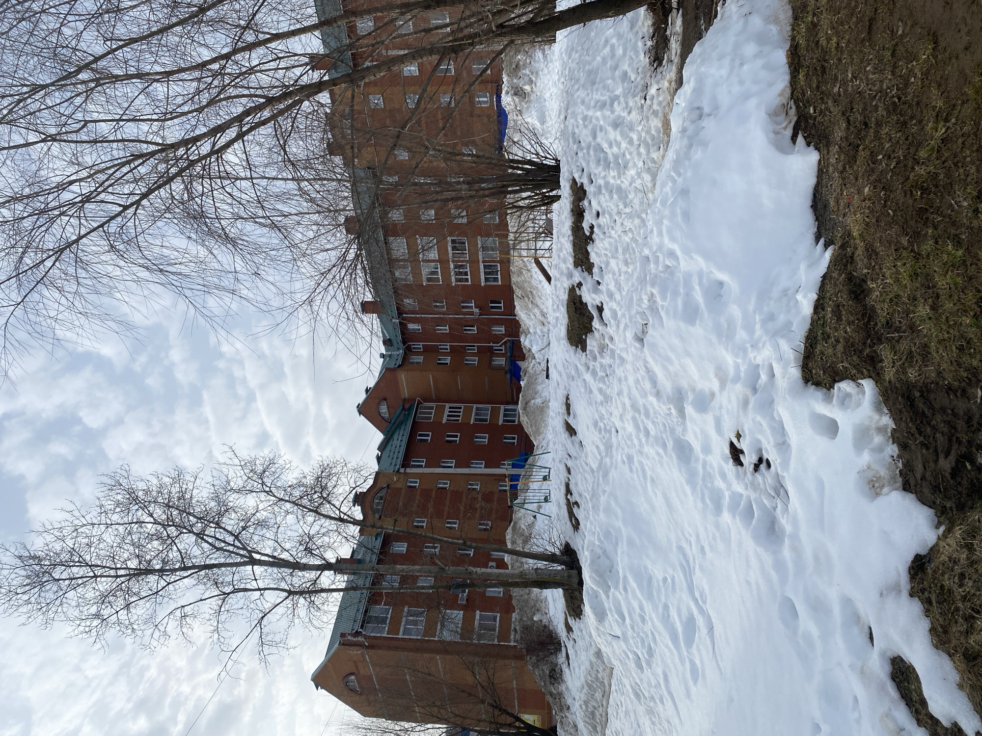 The scene portrays an outdoor area in Chaikovsky, Russia, characterized by late winter or early spring conditions. The ground in the foreground and mid-ground is covered by uneven, melting snow patches, revealing dark brown soil and sparse, dry grass in various areas. Footprints and irregular depressions are visible in the snow.
In the background, a substantial multi-story apartment complex made of reddish-brown brick dominates the view. The building features numerous windows, some with white frames, and distinct green sloped roofs with gables and arched windows on the top floor. Chimneys are visible on the rooftops. The complex appears to have multiple sections, forming an angular layout.
In the mid-ground, a children's playground structure is present amidst the snow, featuring a blue and yellow metal swing set with two swings. Other playground elements are partially obscured by snow. Several bare deciduous trees with thin branches, devoid of foliage, are scattered throughout the image, with some framing the apartment building against an overcast sky filled with white and grey clouds. Faint utility lines are visible in the upper left portion of the sky. No individuals are depicted in the scene, and no specific actions or interactions are occurring.