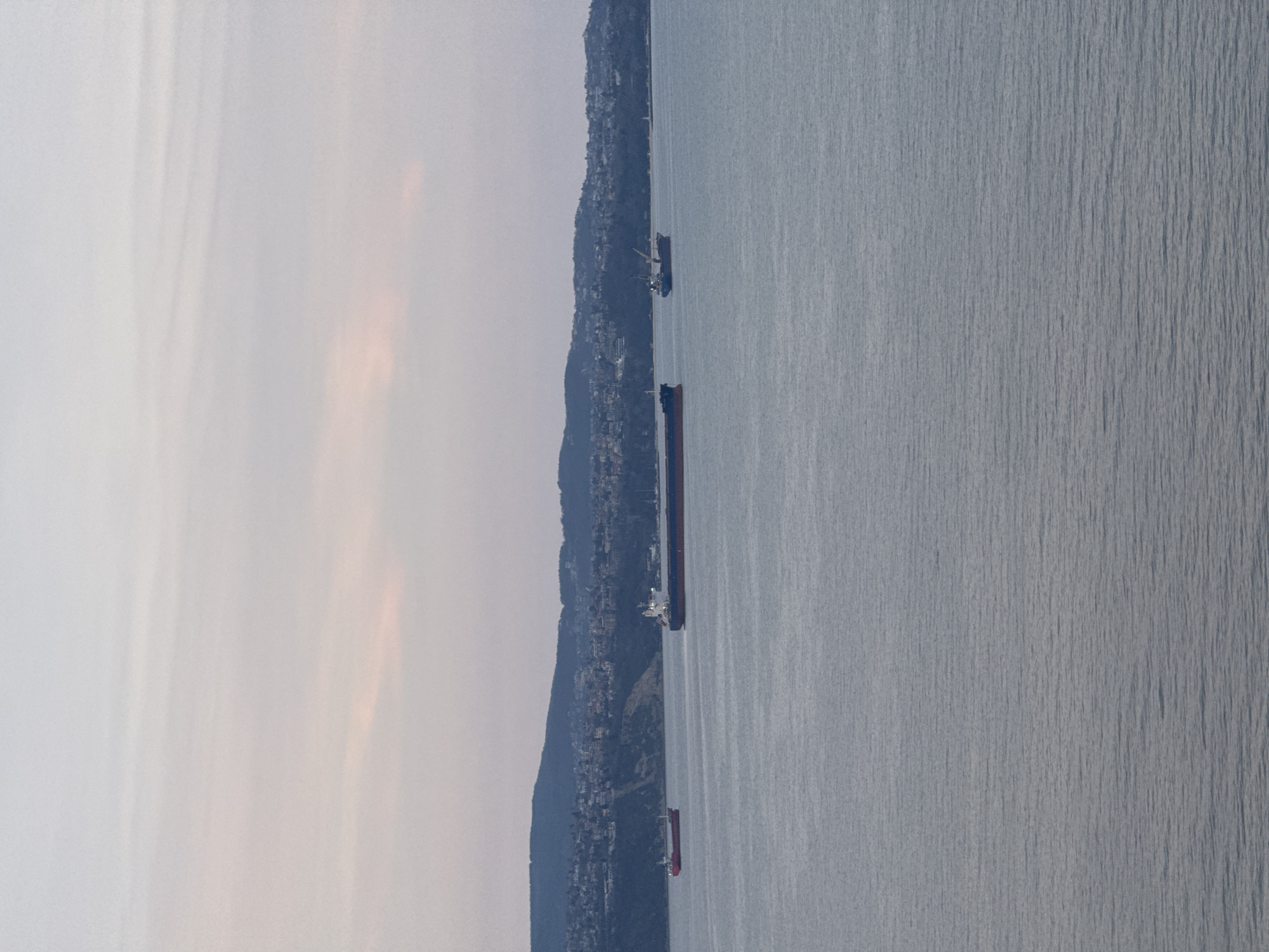 The image shows a view of the Black Sea coastline in Varna, Bulgaria. Along the shoreline, there is a populated hillside with buildings. Several ships are visible in the water, including a larger ship with a red hull. The sky is overcast.