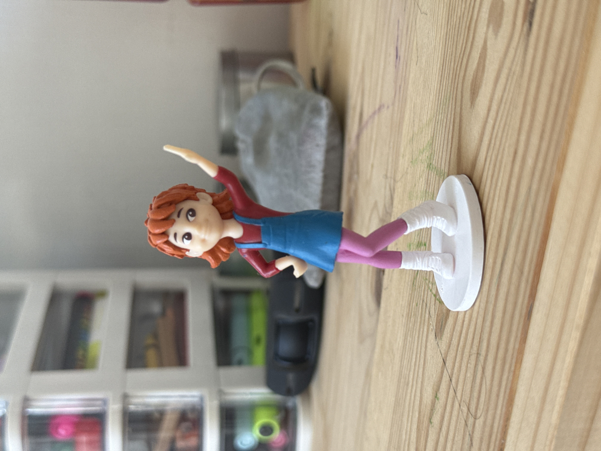 A plastic figurine of a young girl with short orange hair, wearing a red long-sleeved shirt, a blue overall dress, pink tights, and white boots, stands on a white circular base. The figurine is posed with its right arm raised and its left hand on its hip. It is situated on a light brown wooden surface with visible grain and some green and purple markings. In the blurred background, a white multi-drawer cabinet filled with various items is visible to the left, and a large grey fabric object with a metallic handle, possibly a bag or a tea cozy, sits to the right. The scene is illuminated by natural light. There is no indication of the location being in Lyubertsy, Russia.